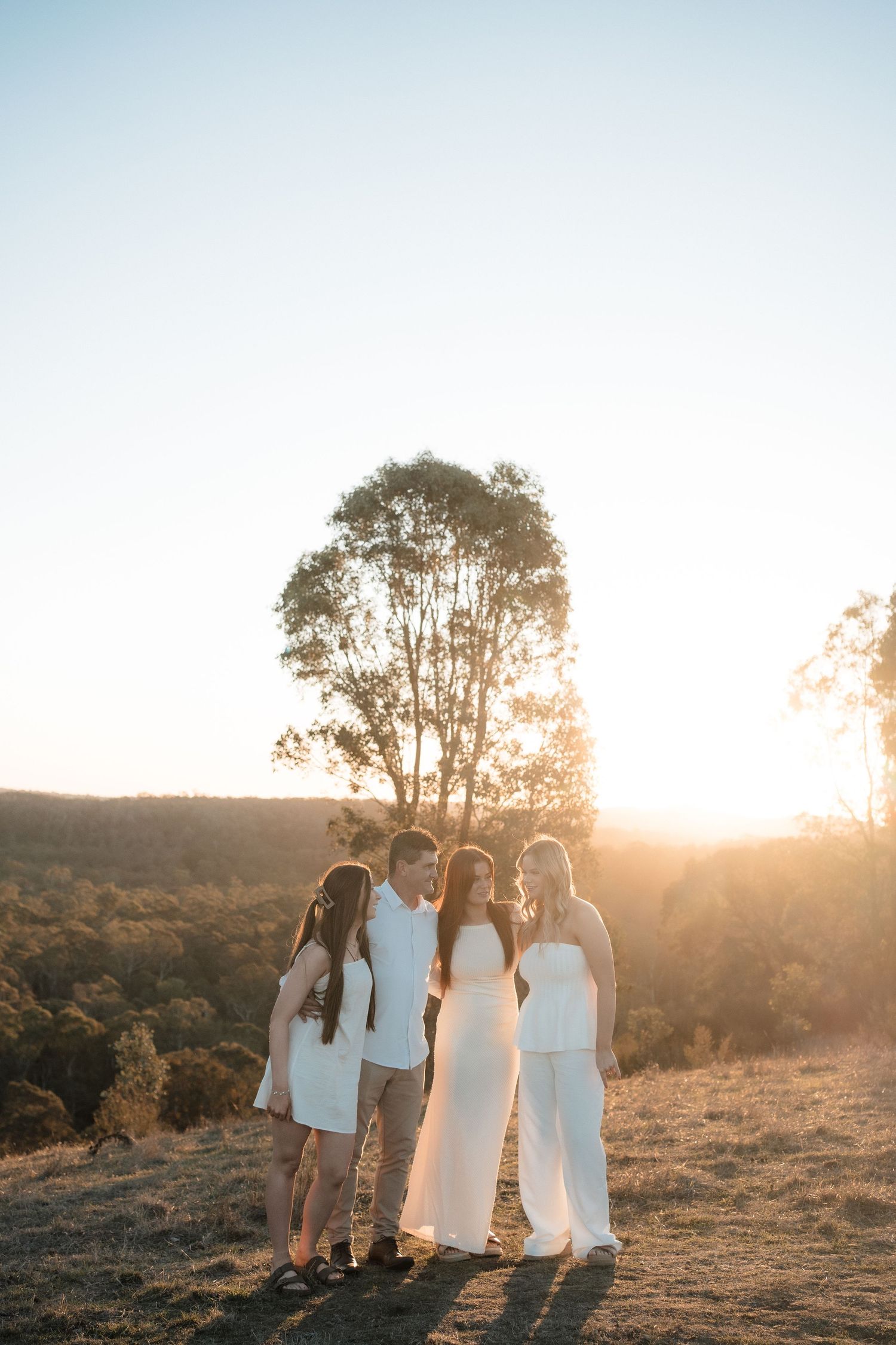 Group in flowy white dresses enjoy golden sunset on a hilltop as warm light filters through trees in background.