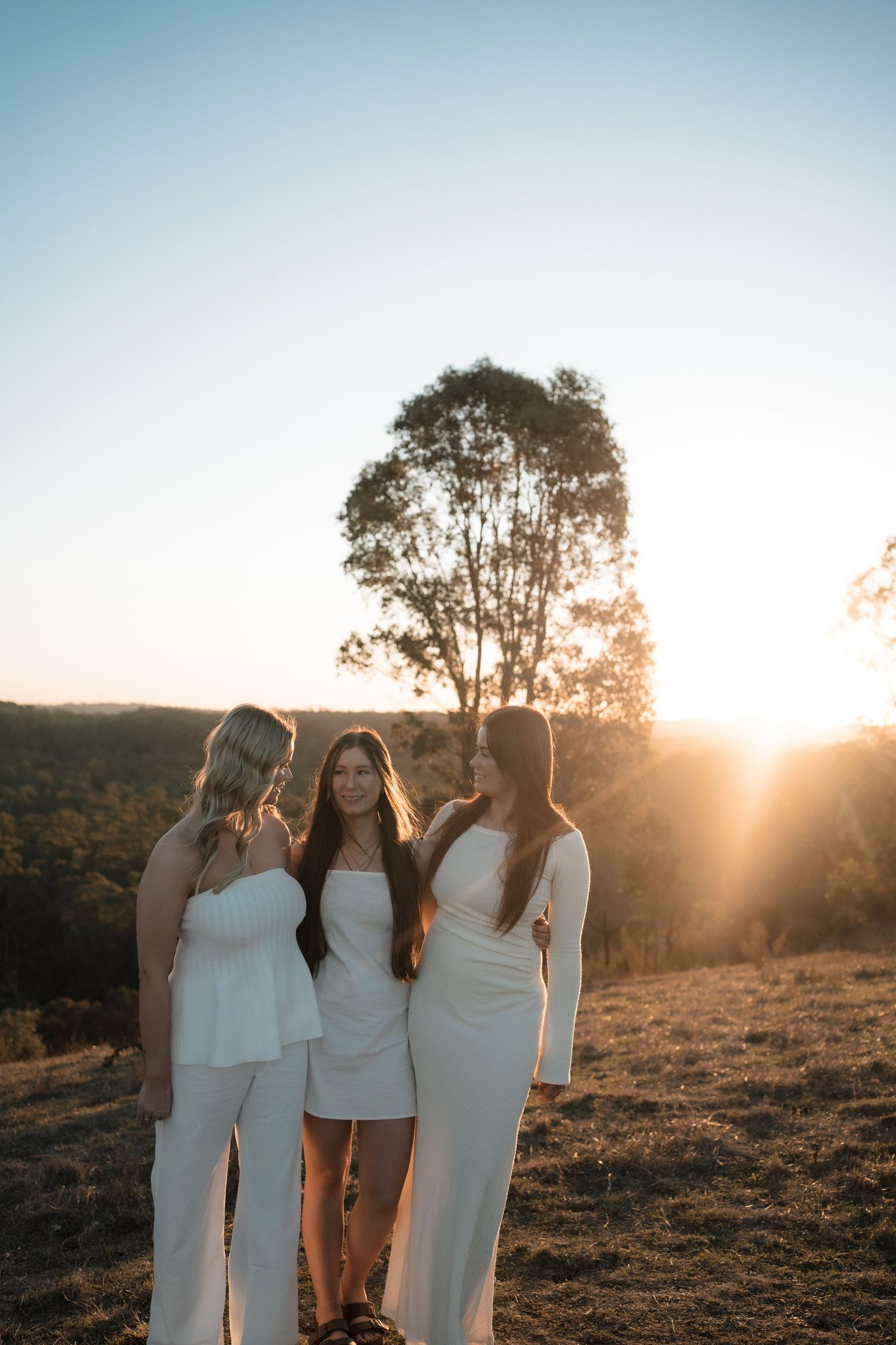 Three figures in white dresses stand together at sunset with a tall tree silhouetted against the golden sky.