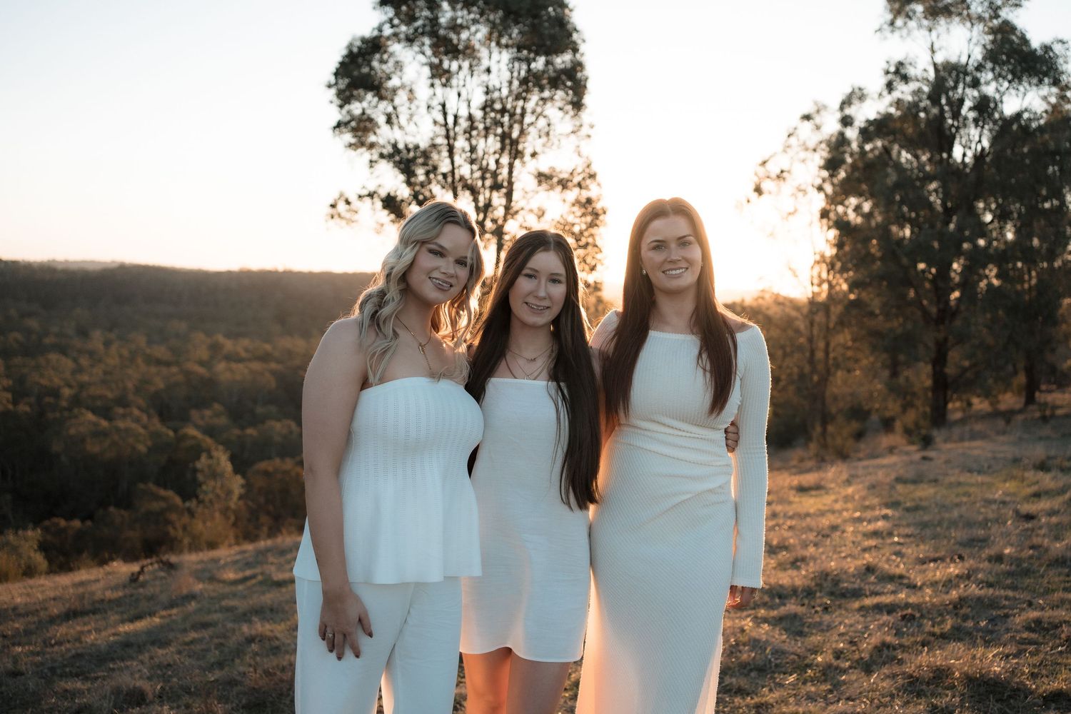 Three friends in white dresses pose together on a hilltop at sunset with trees and mountains in the background.