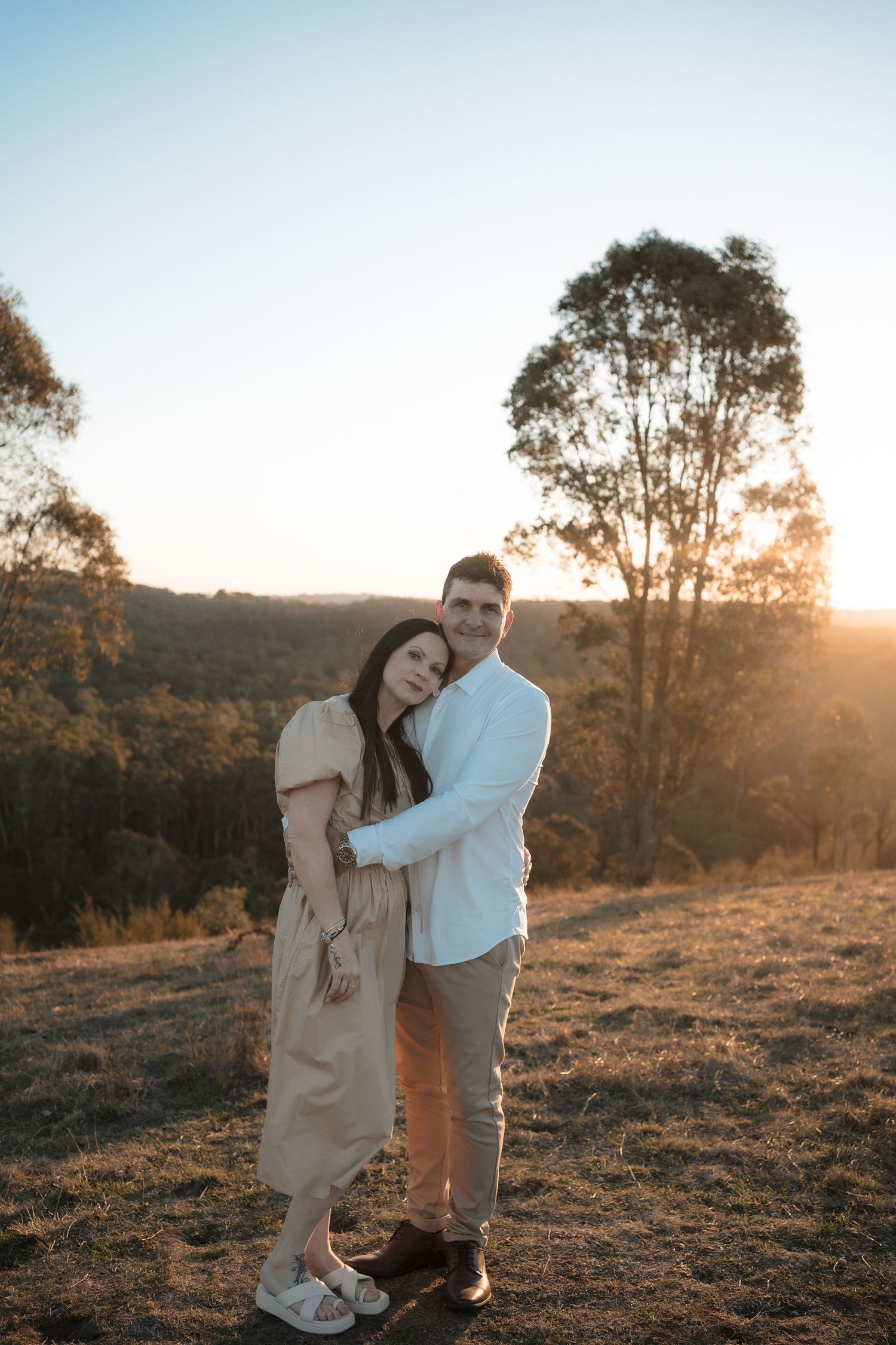 A couple embraces during golden hour sunset in a rural setting with eucalyptus trees silhouetted in the background.