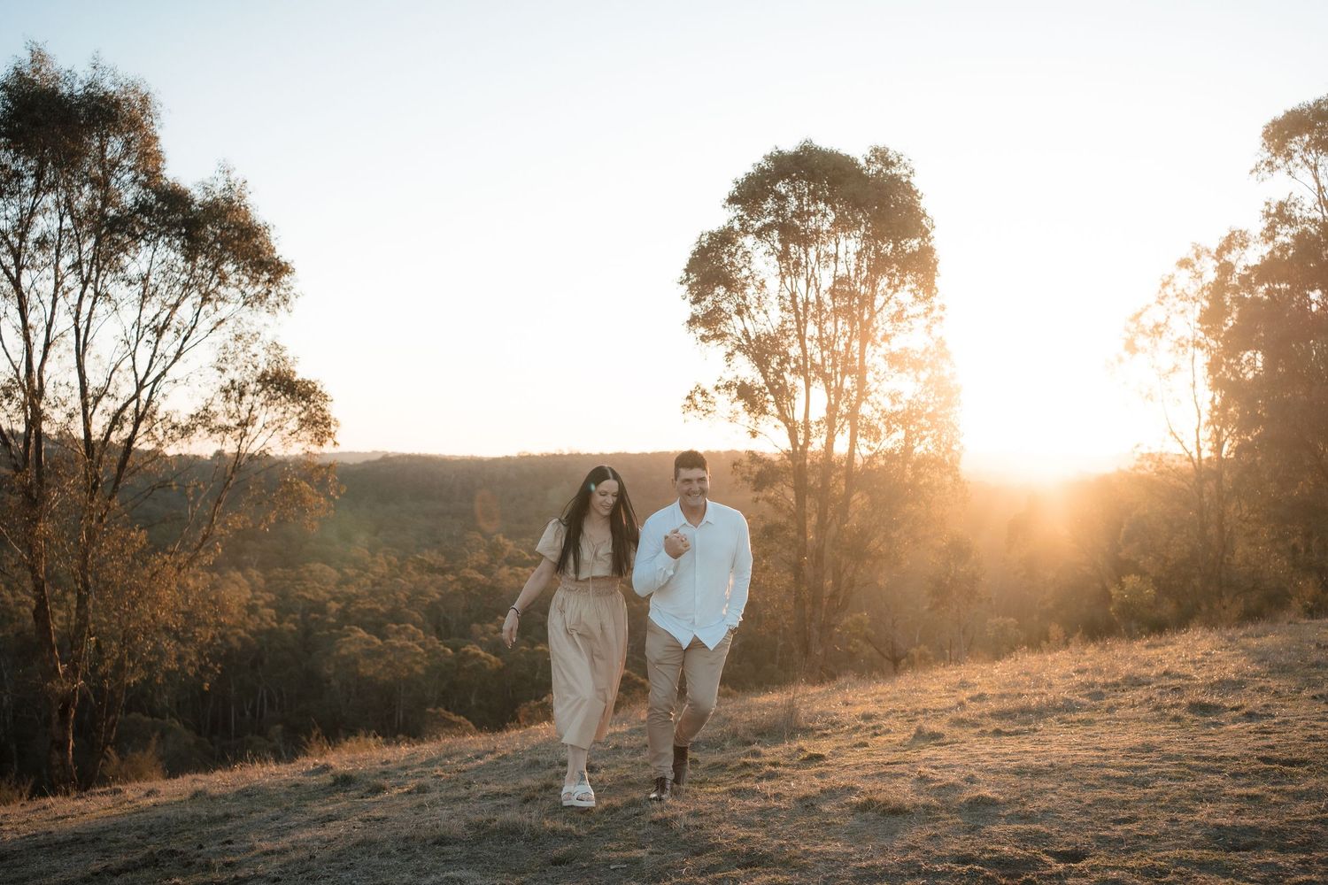 Two people walk hand in hand on a hillside path during golden sunset with eucalyptus trees silhouetted in background.