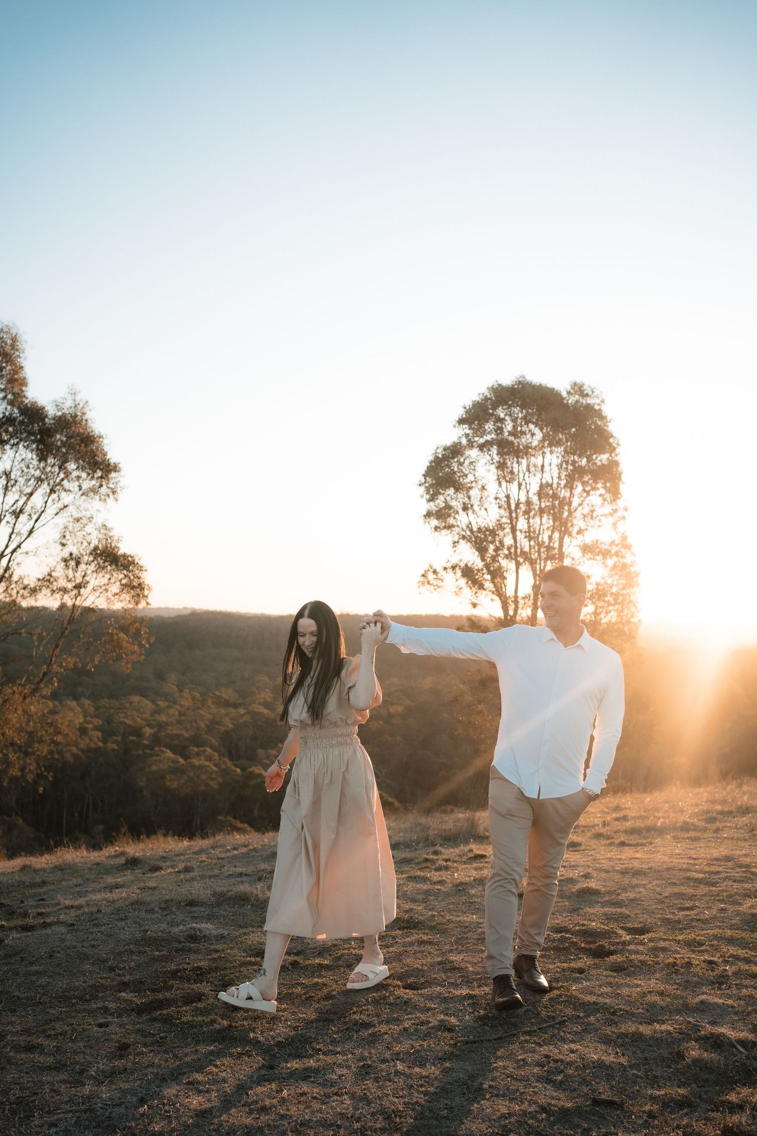 A couple walks together on a hillside during sunset with tall trees casting long shadows in the warm evening light.