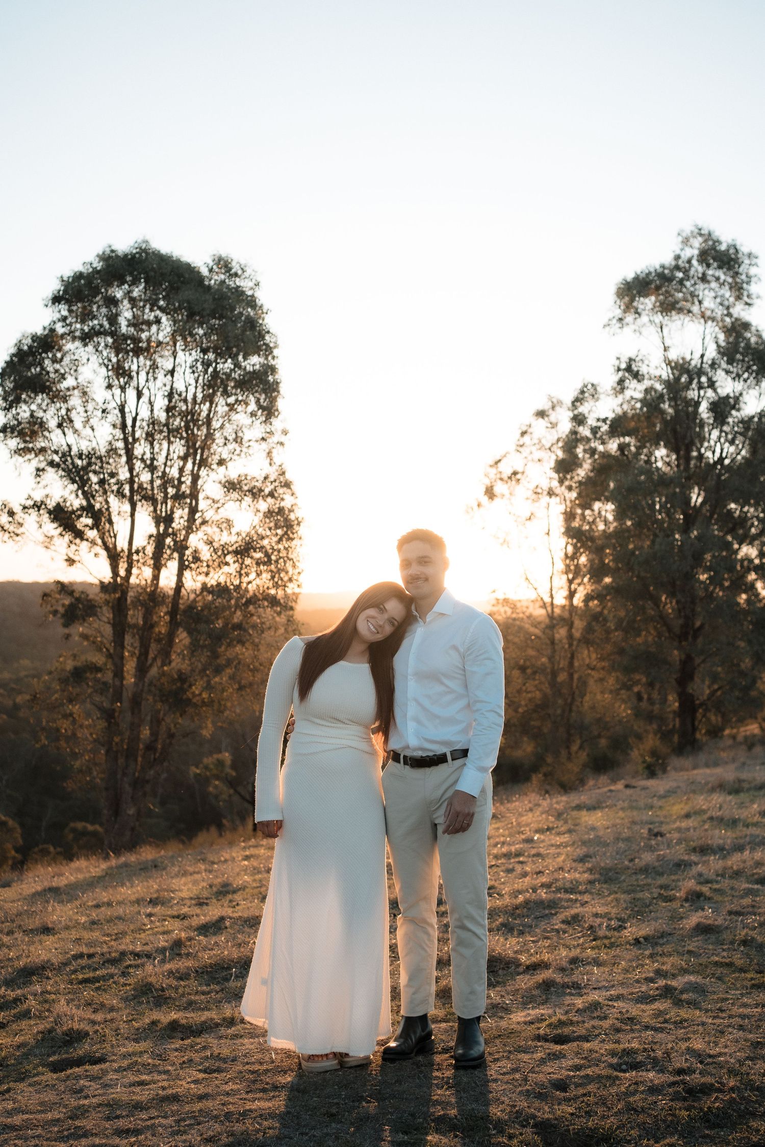 A couple in white outfits stands together on a hillside during golden hour sunset with trees silhouetted in the background.