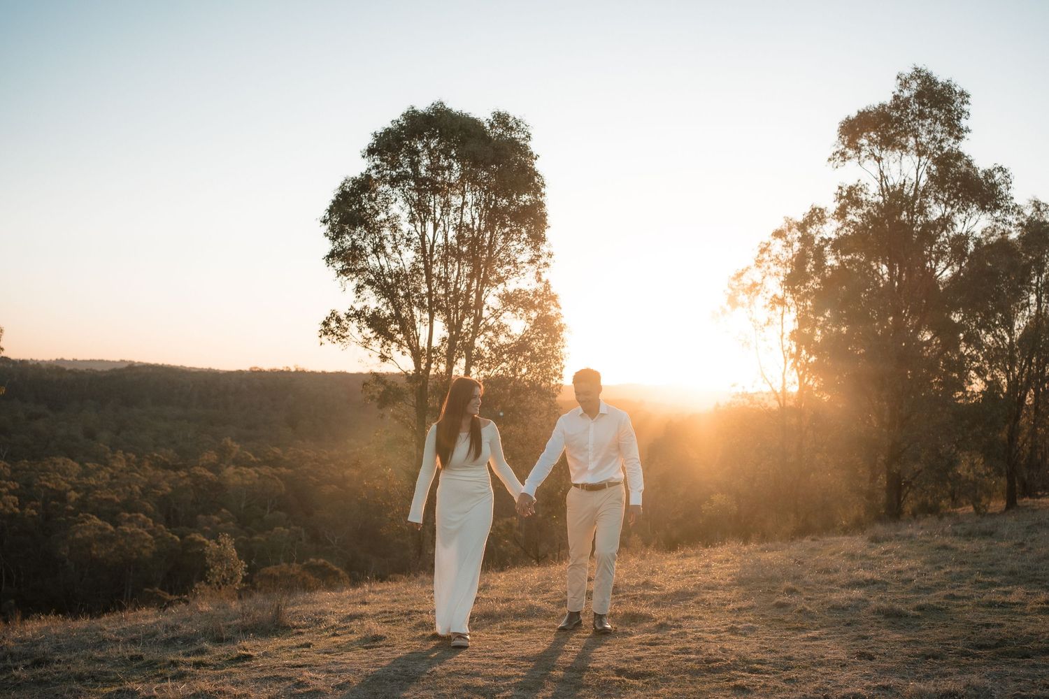 Two figures in white outfits walk hand in hand on a hilltop at sunset with tall eucalyptus trees silhouetted against the sky.