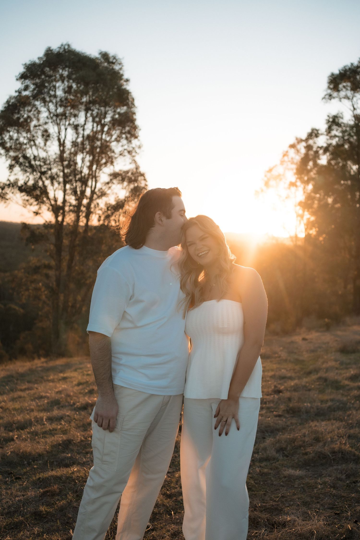 Two people dressed in all white embrace on a grassy slope during sunset with eucalyptus trees in the distance.