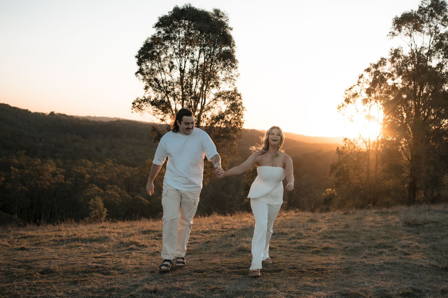A couple walking hand in hand on a hilltop at sunset with eucalyptus trees silhouetted in the golden light.