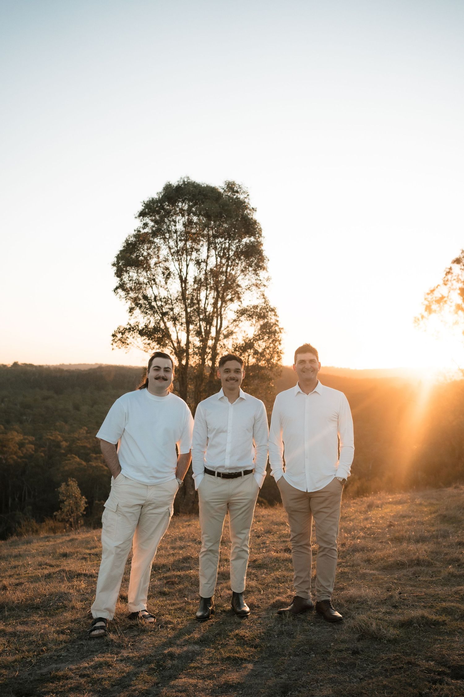 Three people stand together on a hilltop during golden hour sunset with a large tree silhouetted in the background.