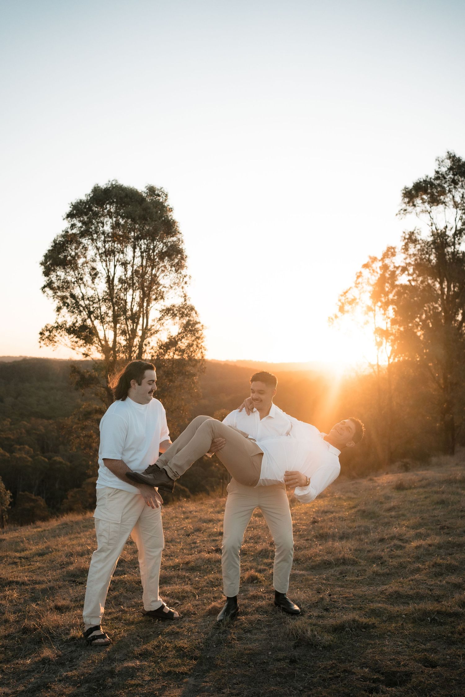 Two figures in white outfits stand together against a sunset on a grassy hillside with silhouetted trees.
