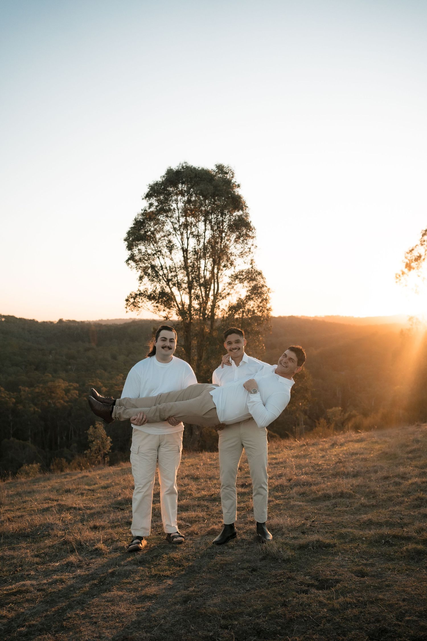 Three people in white outfits pose playfully on a hilltop at sunset with a silhouetted tree in the background.