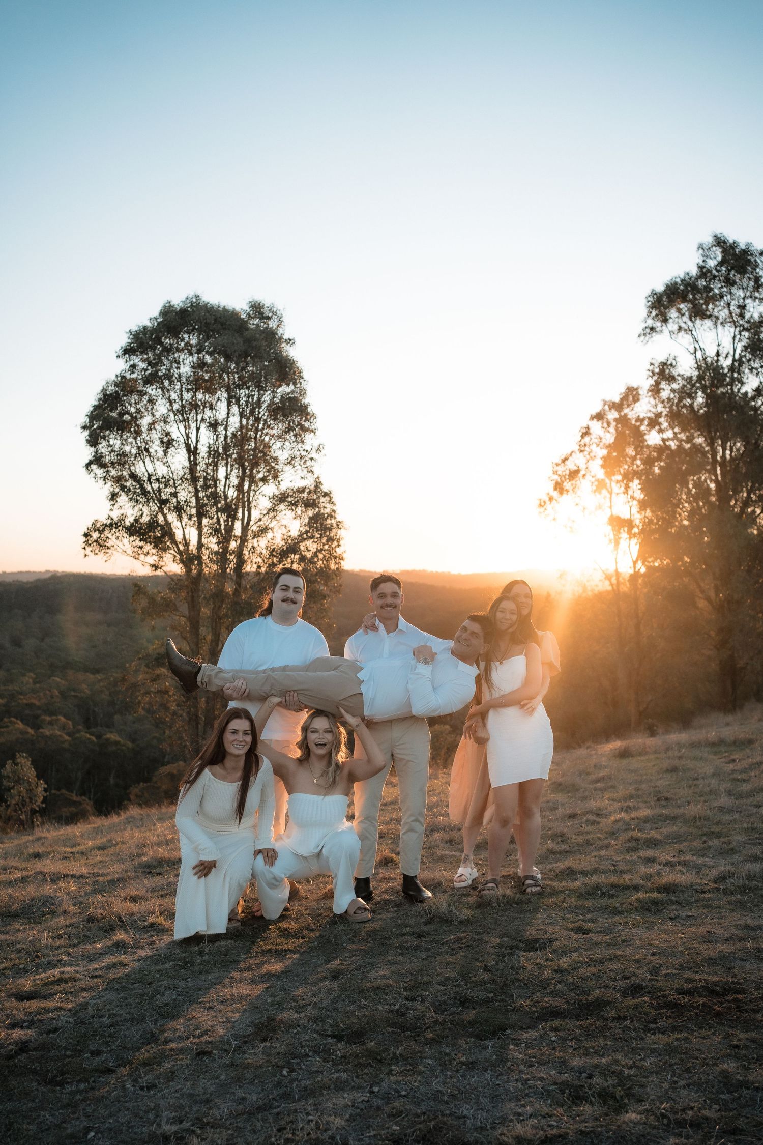 Group wearing white outfits poses together on a hilltop during golden hour sunset with eucalyptus trees in background.