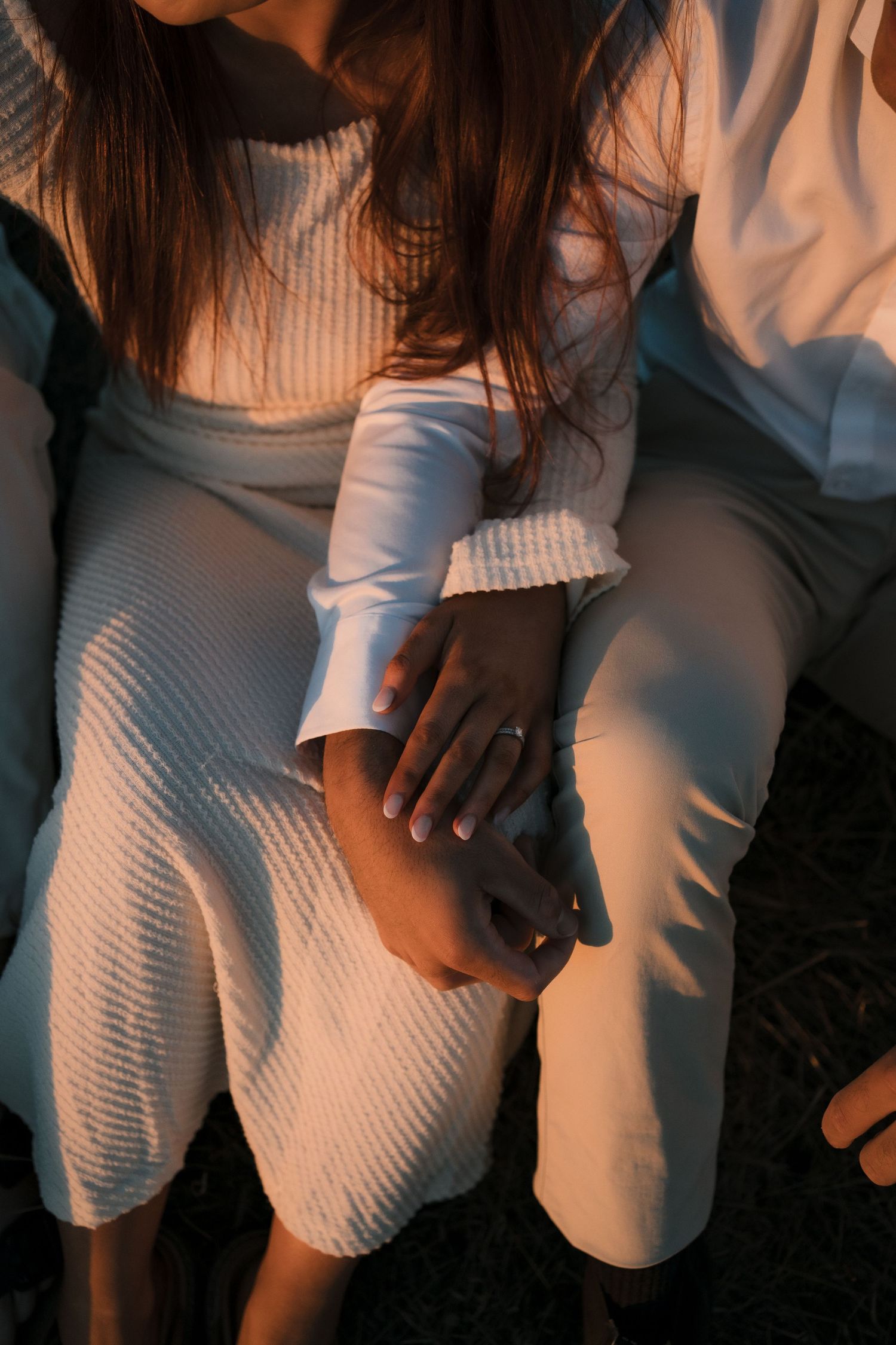 Person wearing white ribbed sweater and pants sitting on dark surface in moody lighting at sunset.