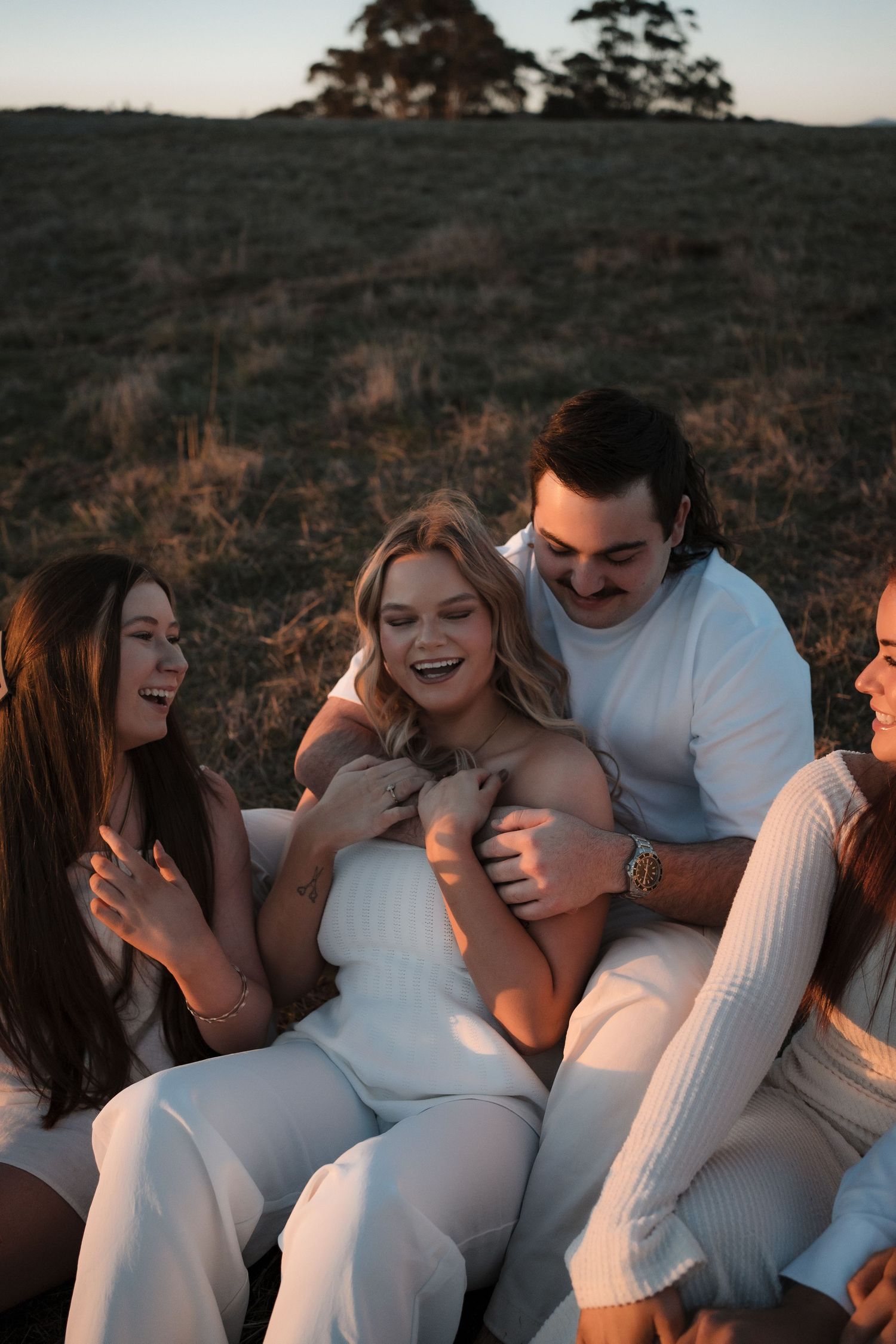 Group of friends in all-white outfits sharing joyful laughter together outdoors during golden hour.