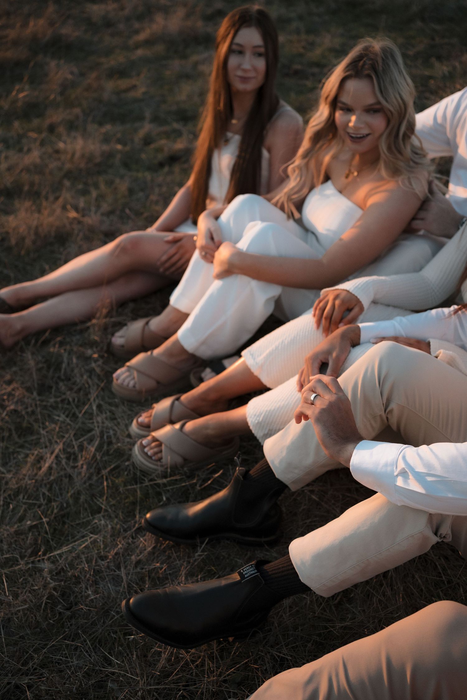 Group relaxing on grass hillside at sunset wearing white and neutral outfits with boots.