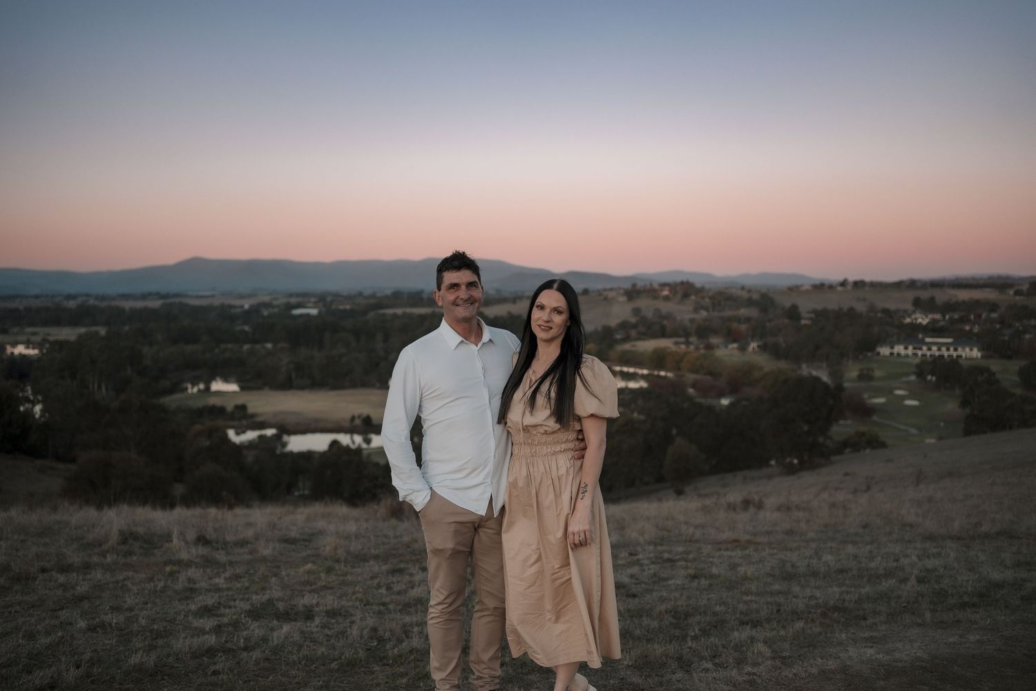 A couple poses together on a hilltop overlooking a rural valley landscape at dusk with soft pink sky.