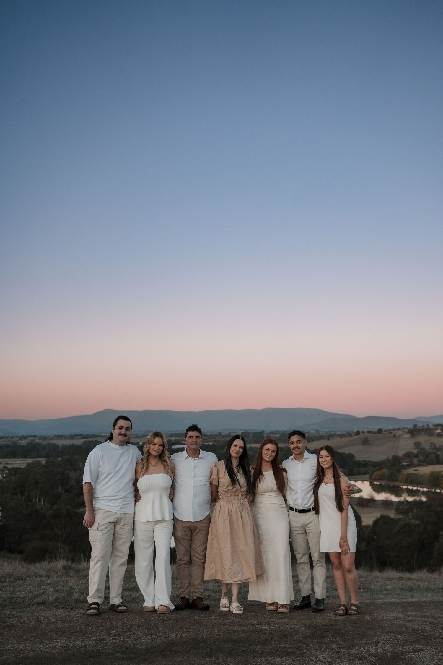 Group portrait of friends wearing coordinated white and neutral outfits against a scenic mountain backdrop at dusk.