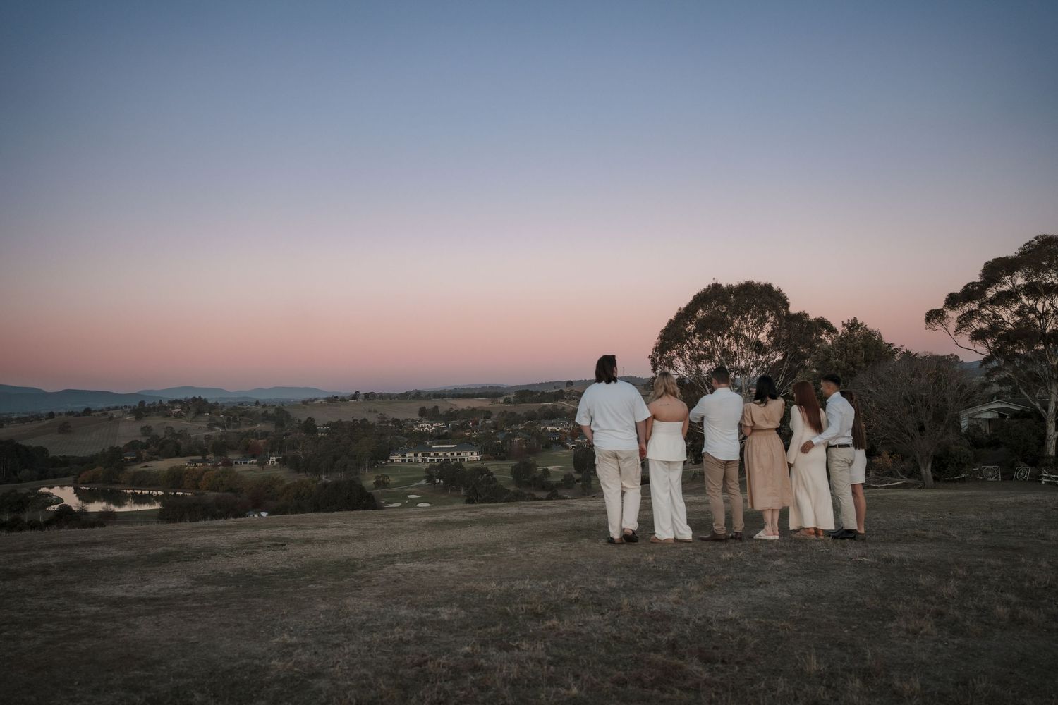 A family of four stands together watching the sunset over rolling hills in matching white and cream outfits.