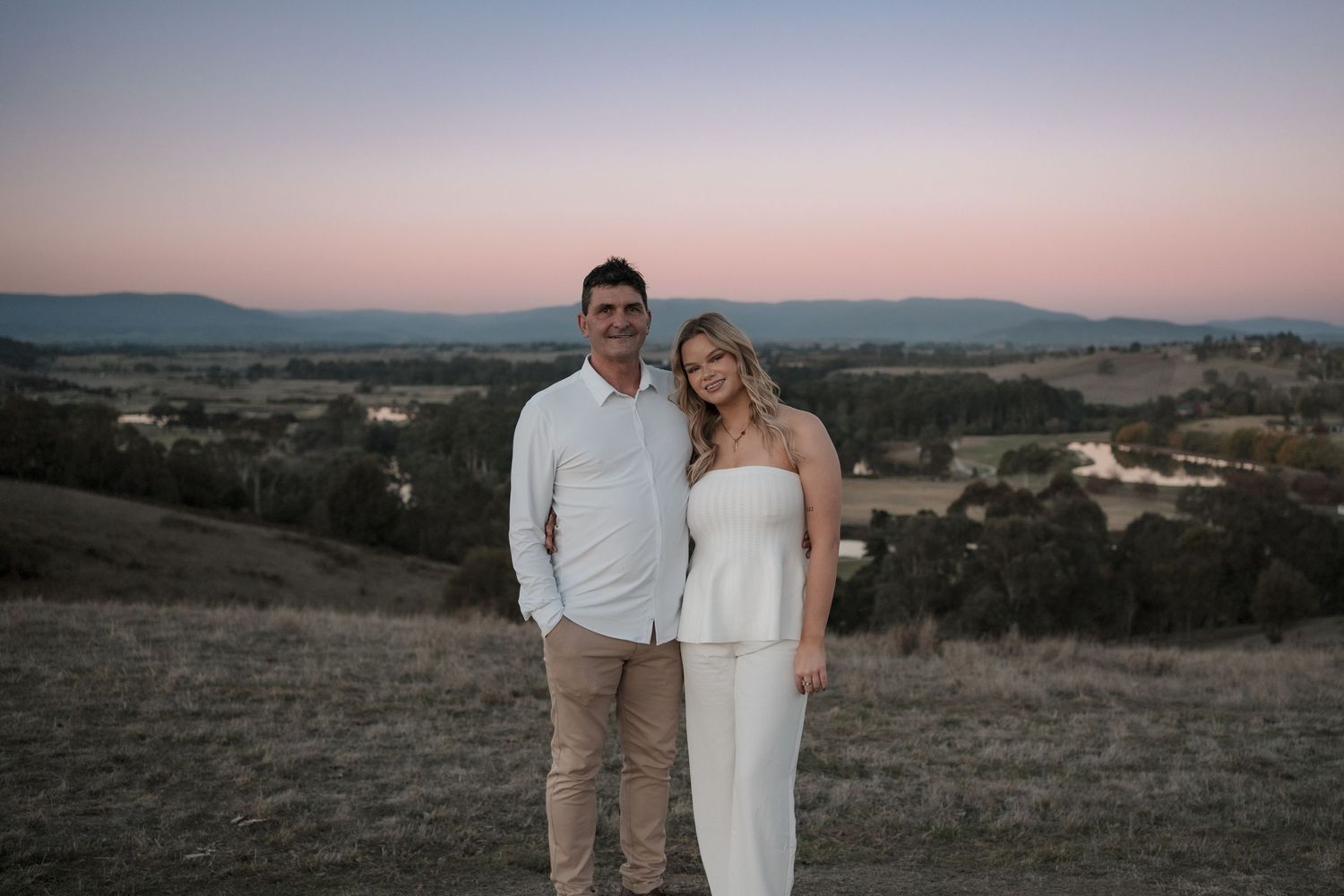 A couple stands together on a hilltop during sunset with rolling hills and mountains in the background.