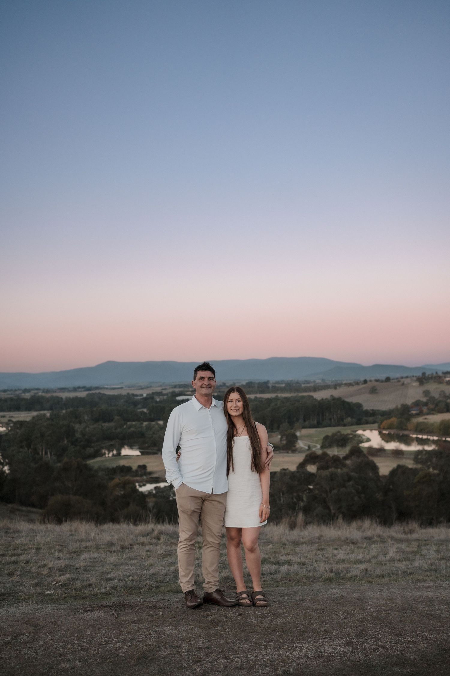 A couple stands together on a hilltop overlooking a scenic valley at sunset with mountains in the distance.