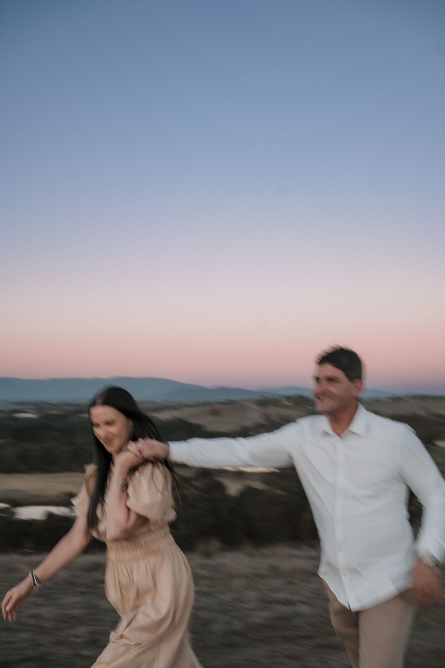 Couple in light-colored outfits enjoying a playful moment against a dusky mountain landscape.