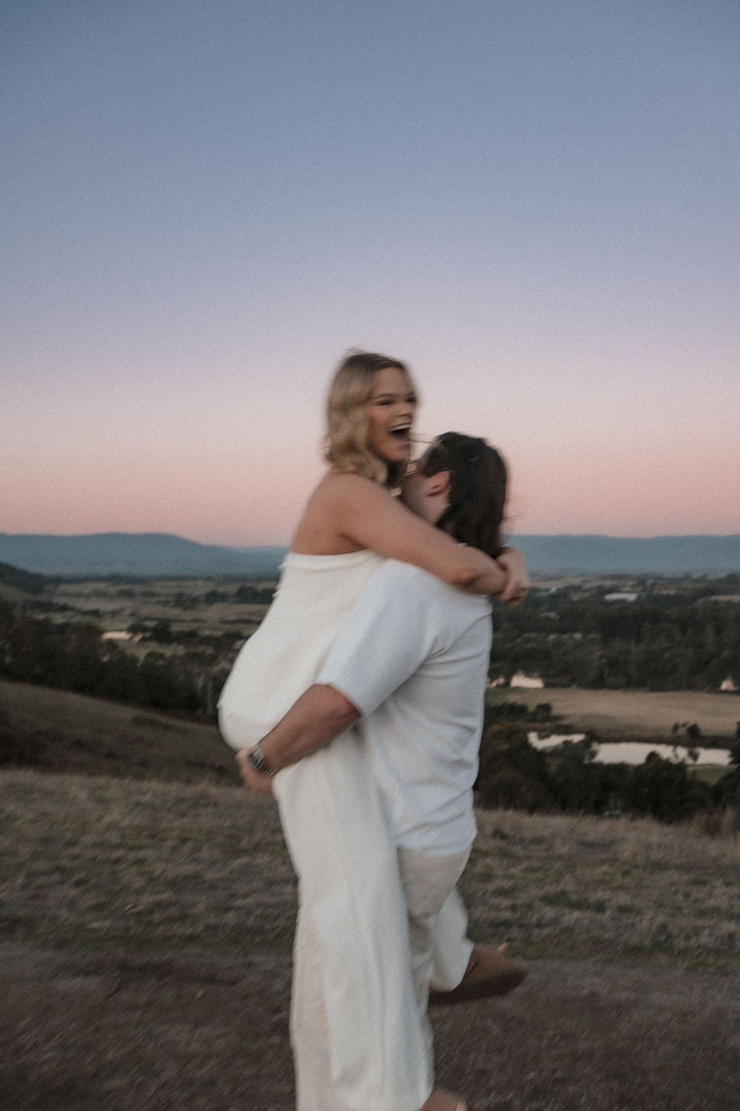 A couple shares a joyful embrace on a hilltop at sunset with rolling countryside and mountains in the distance.