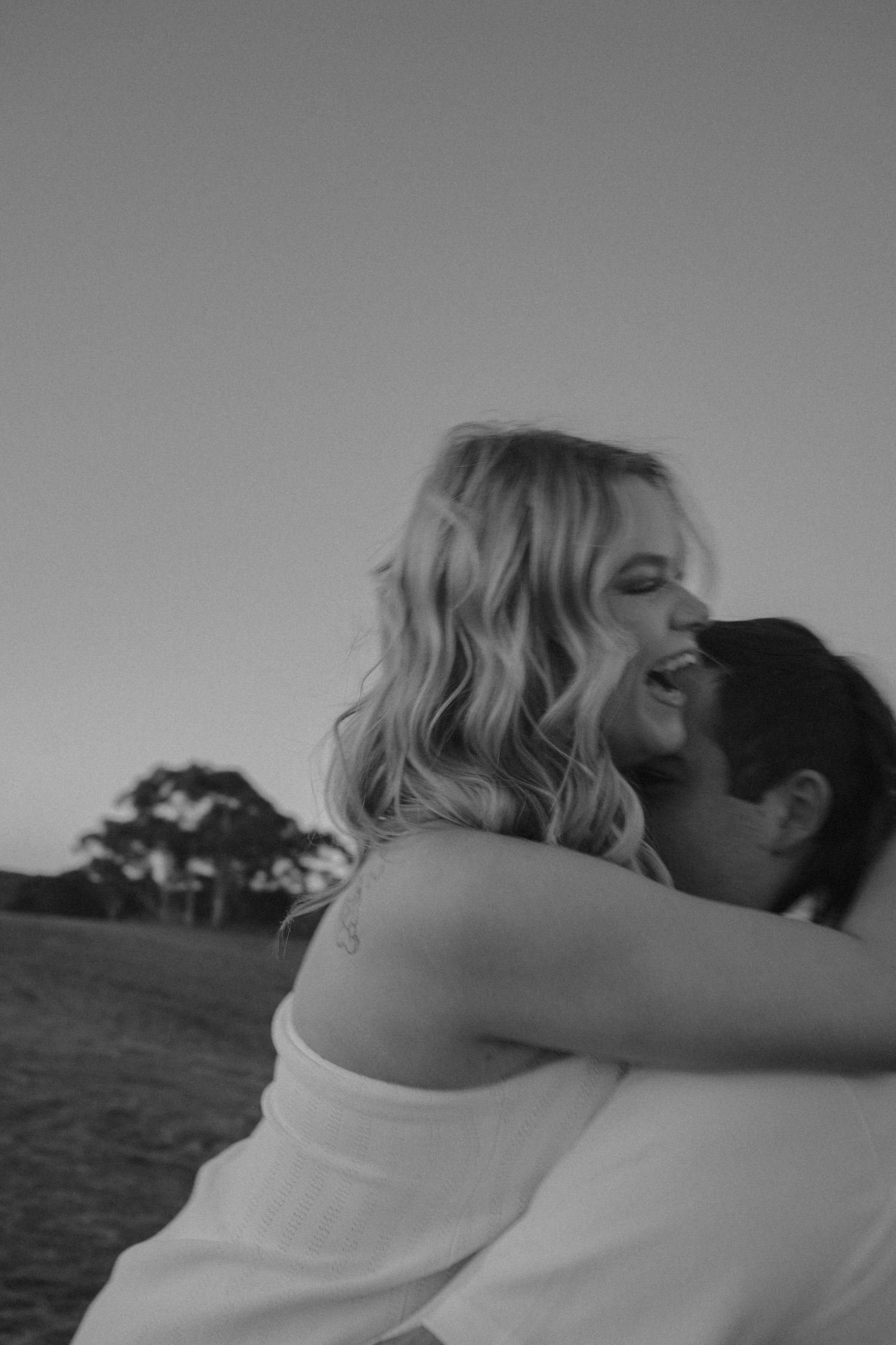 A couple in white embracing outdoors against a blurred natural background in black and white.