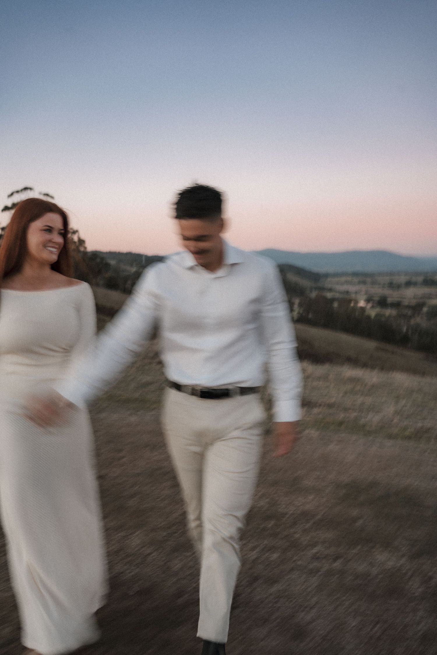 Blurry motion shot of a couple in white outfits walking together on a hilltop at sunset with mountains in background.