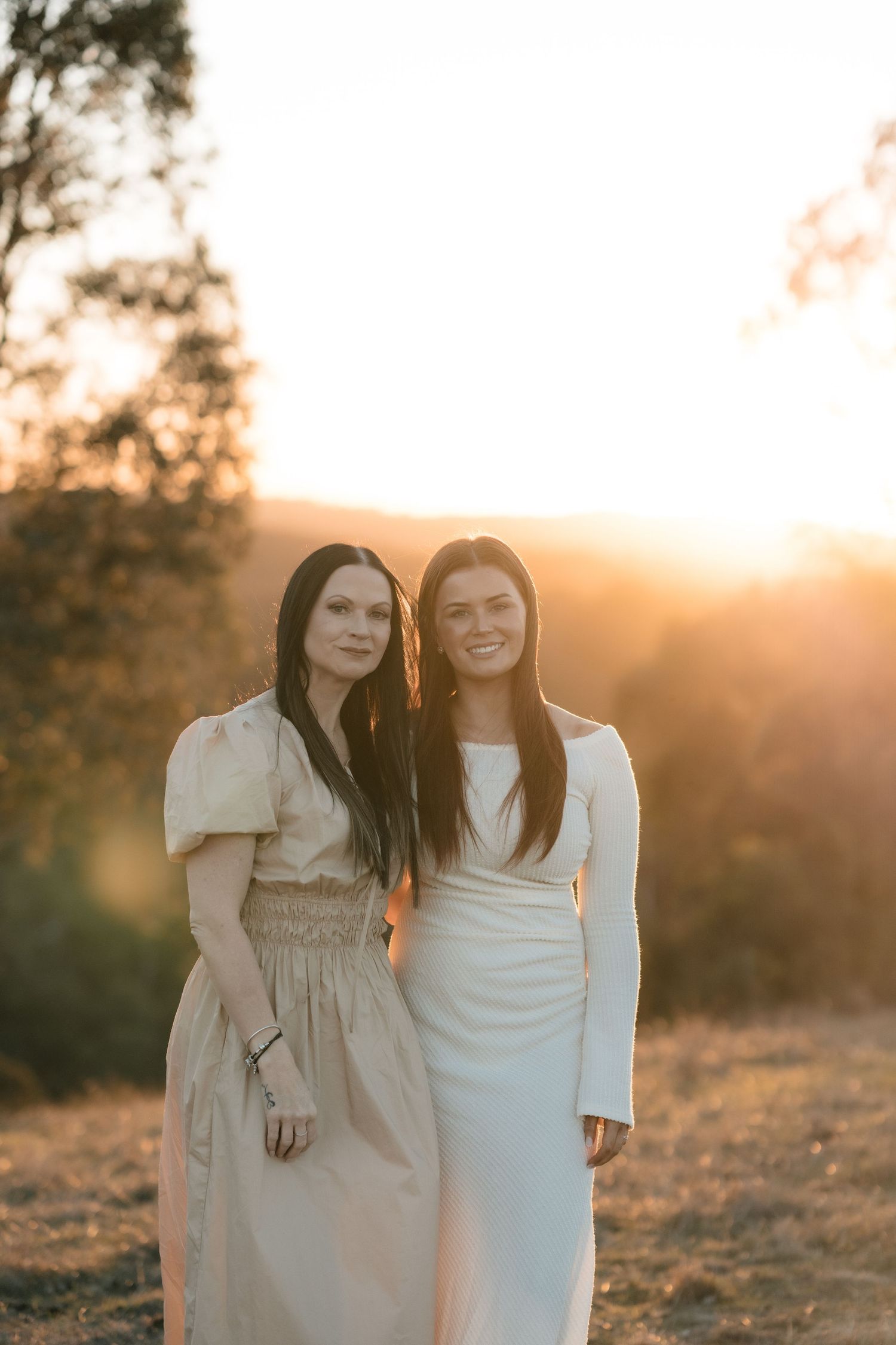 Two people in long dresses stand together outdoors during golden hour sunset in a natural setting.