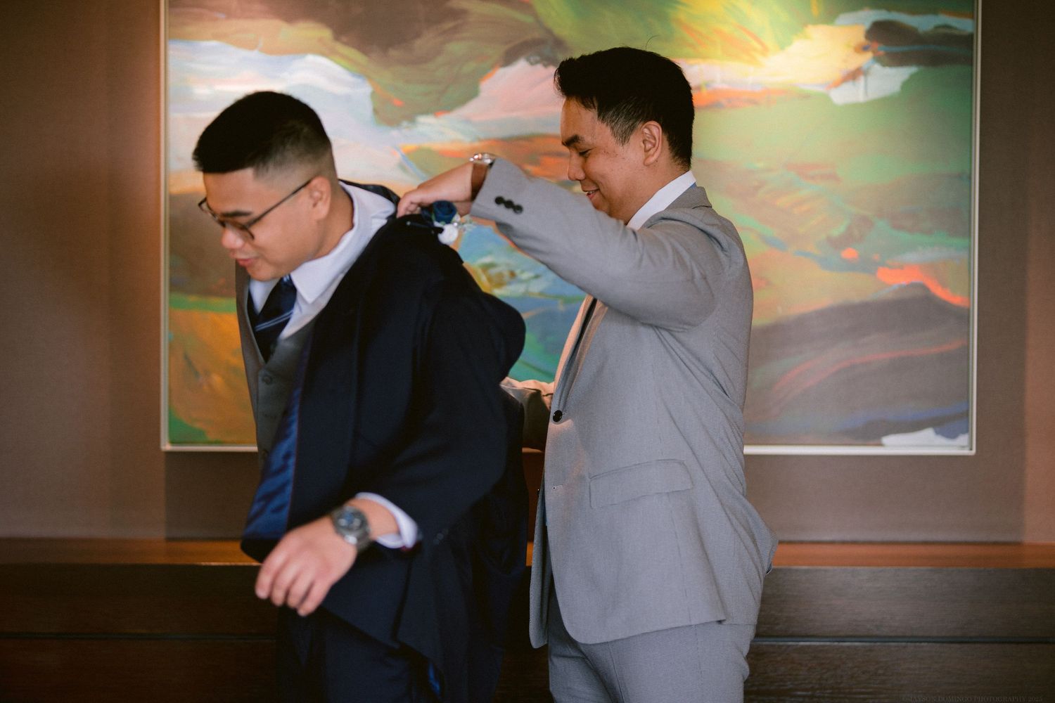 Two people in formal suits assist with wedding preparations in front of a colorful abstract painting.