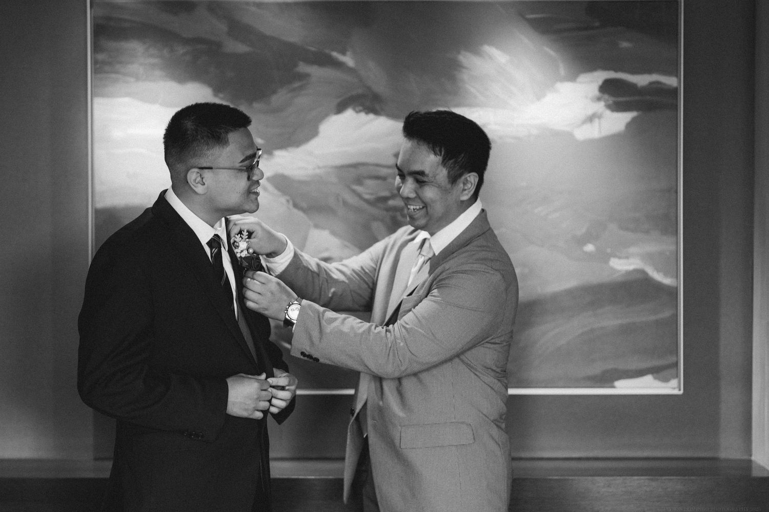 Black and white photo of two men in suits adjusting boutonnieres against painted backdrop.