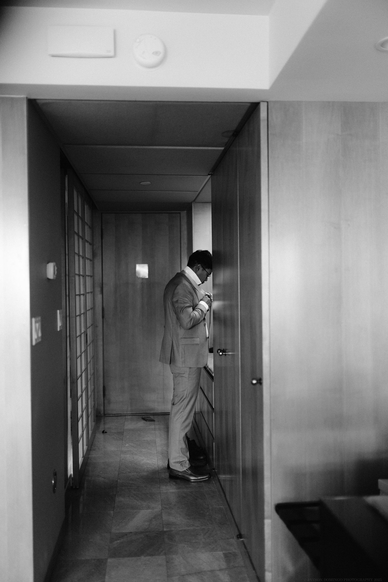 Black and white photo of a suited figure checking phone in a modern hotel hallway.