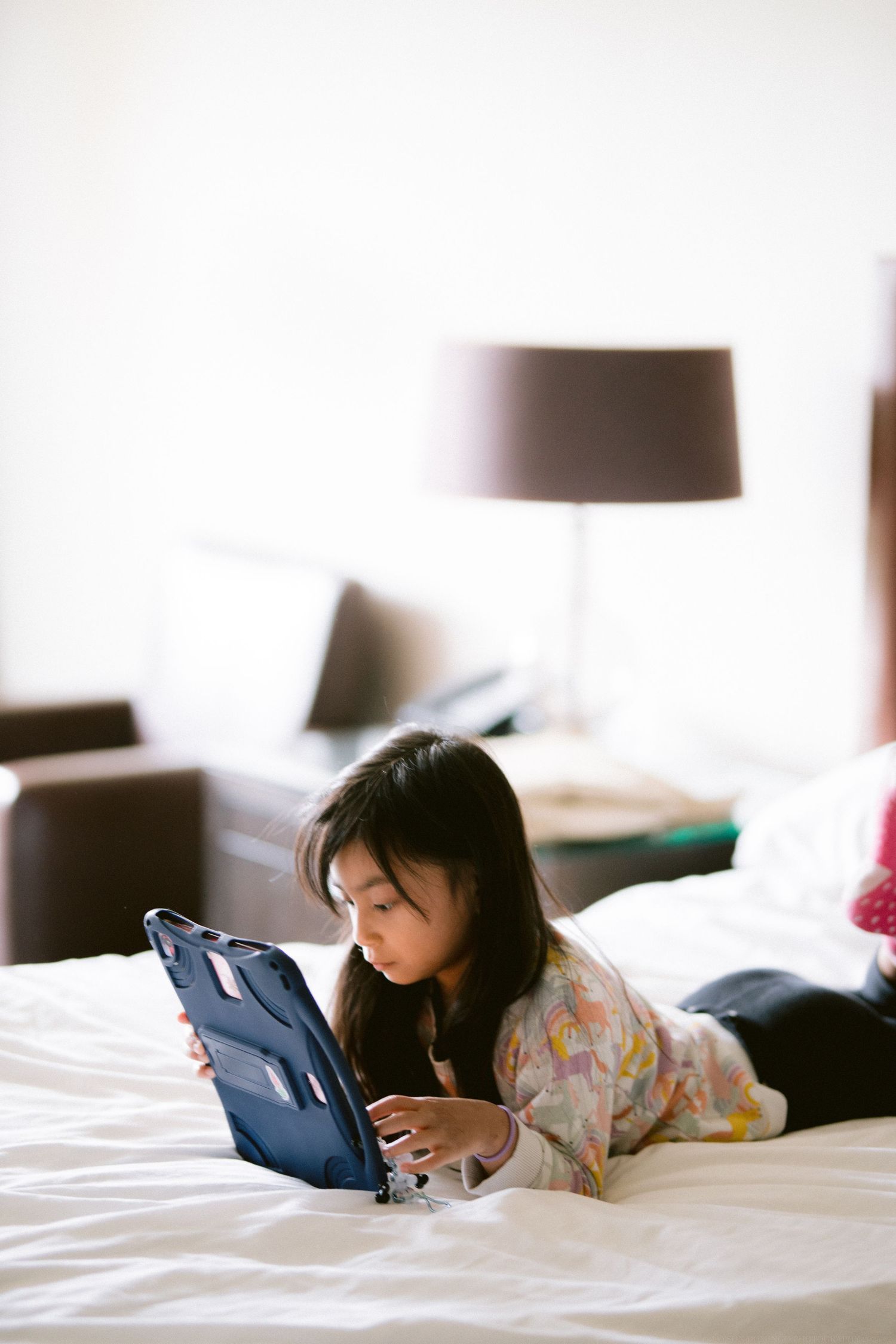 Young child lying on white bedding while using a tablet device in a bright room.
