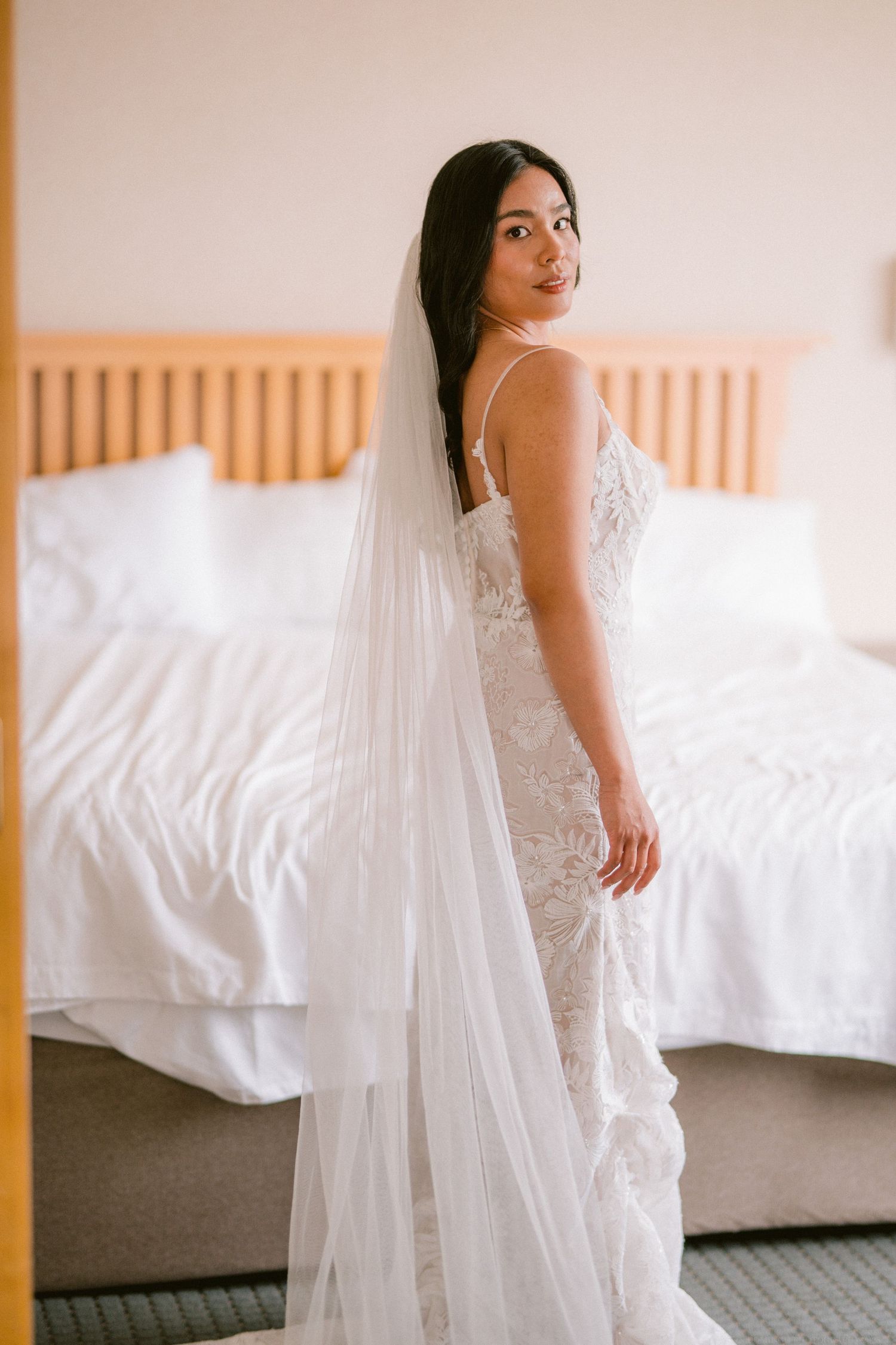 Bride in a lace wedding gown and long veil poses in a soft-lit bedroom.