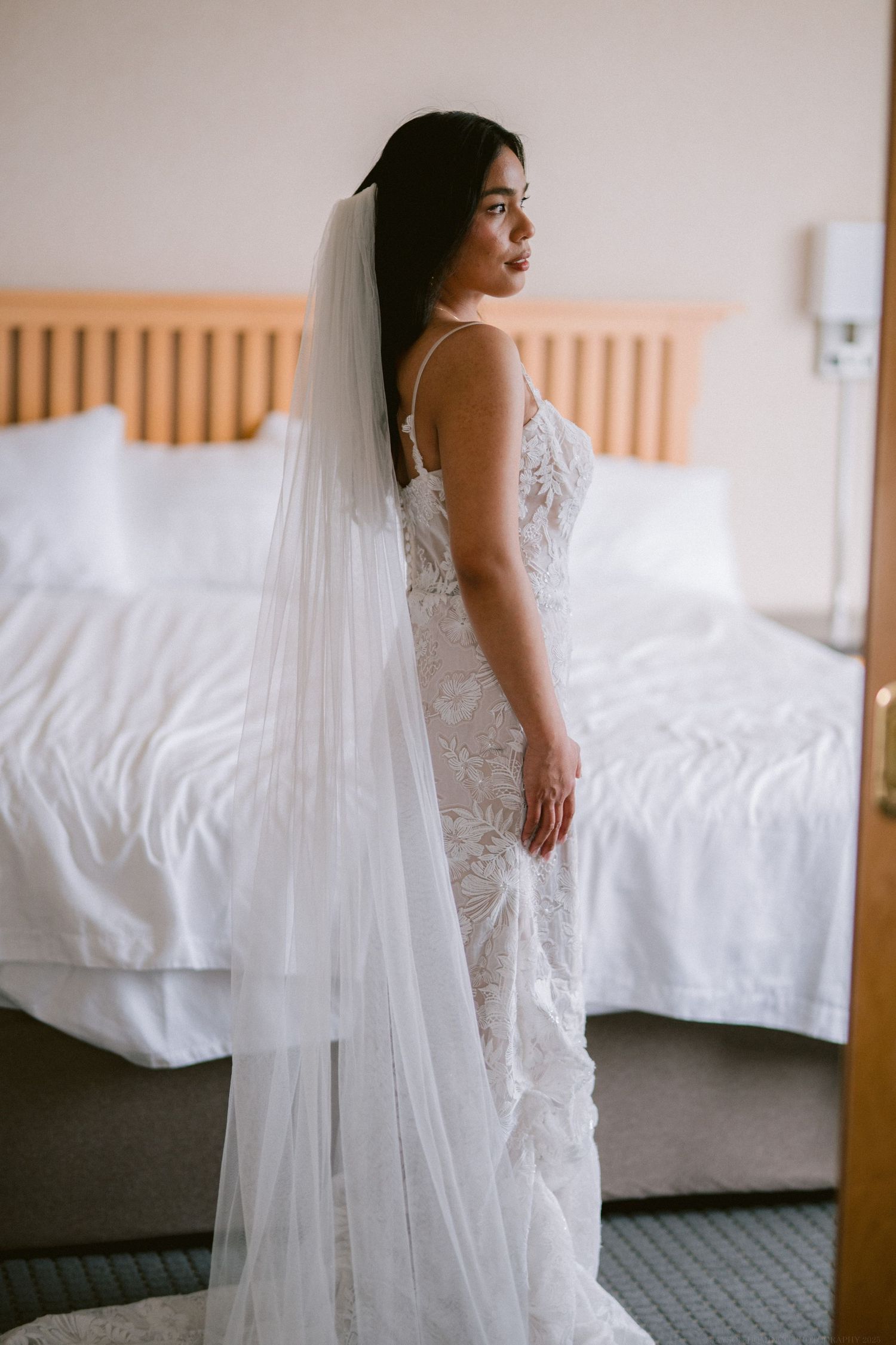 A bride in a lace wedding dress and long veil poses in front of a bed.
