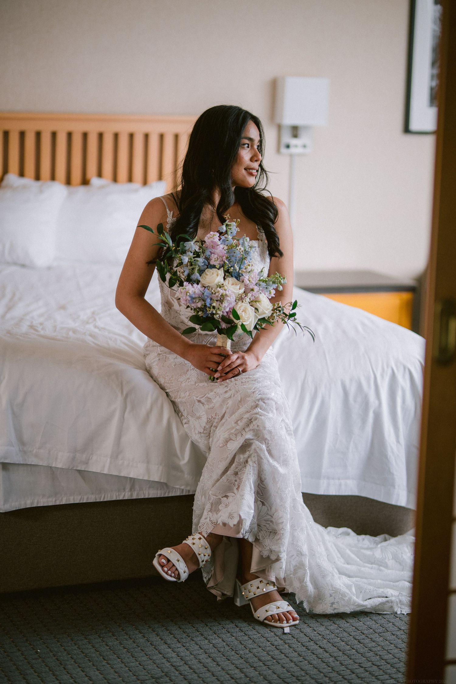 A bride in a lace wedding dress sits on a bed holding a bouquet of pink and purple flowers.