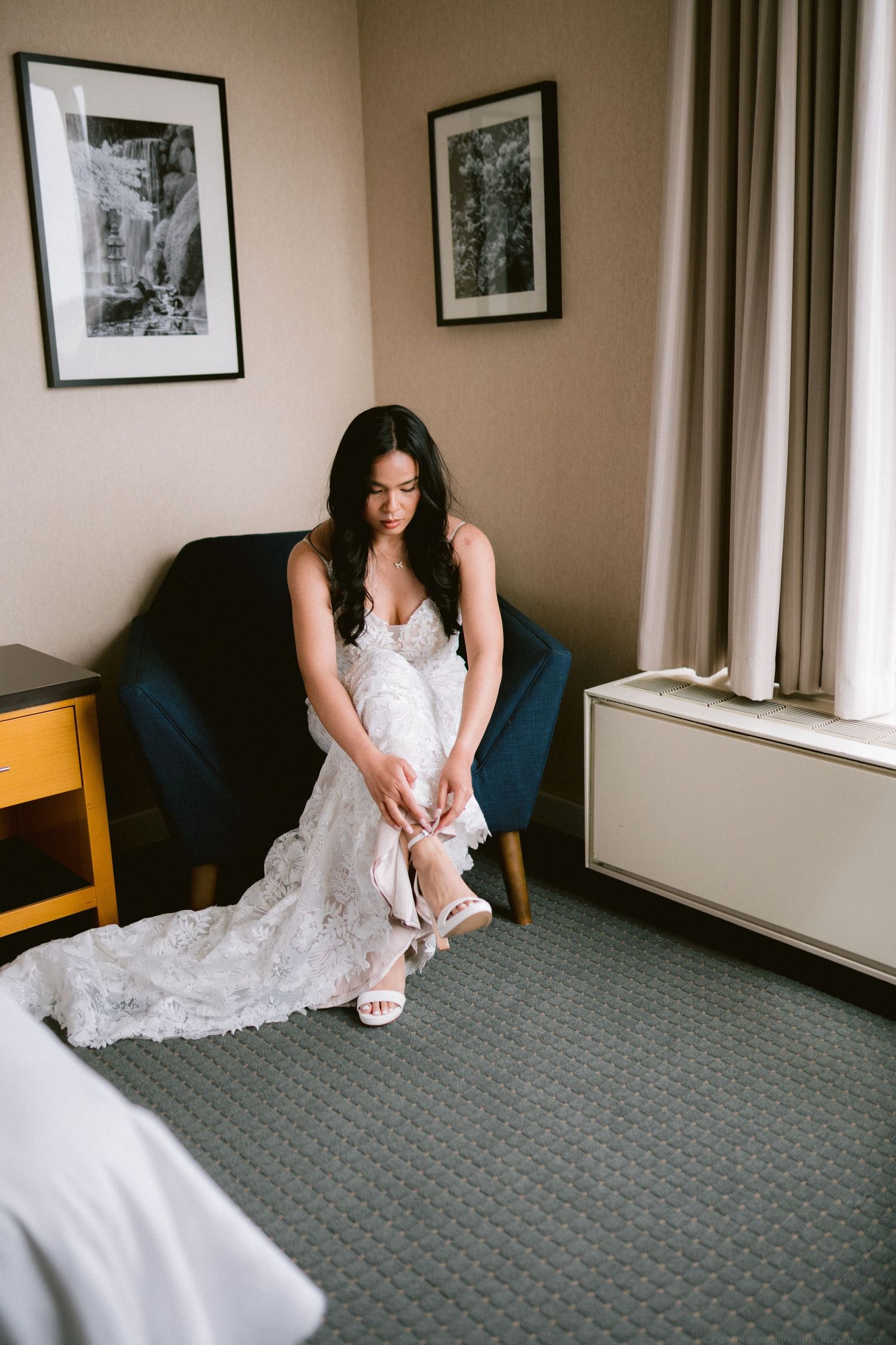 A bride in white lace gown sits on blue chair putting on wedding shoes.