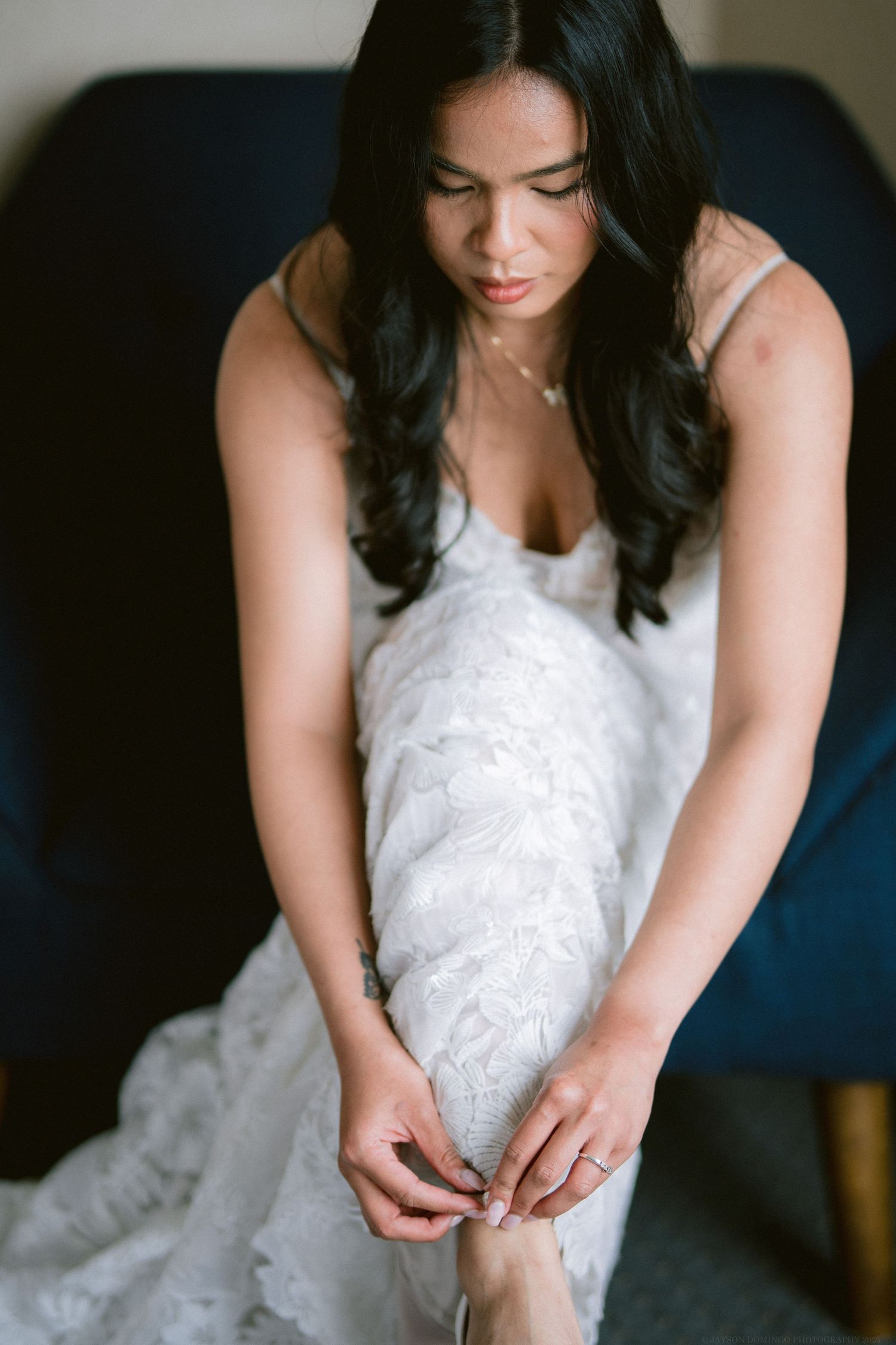A bride in a white lace gown adjusts her shoe while sitting on a blue chair.