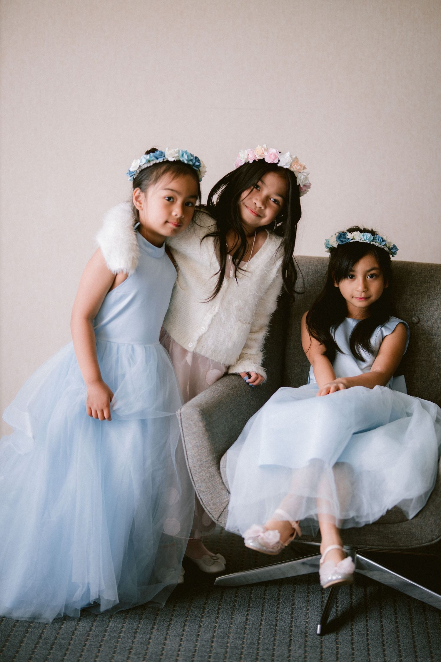 Three flower girls in light blue tulle dresses and floral crowns pose on a couch.