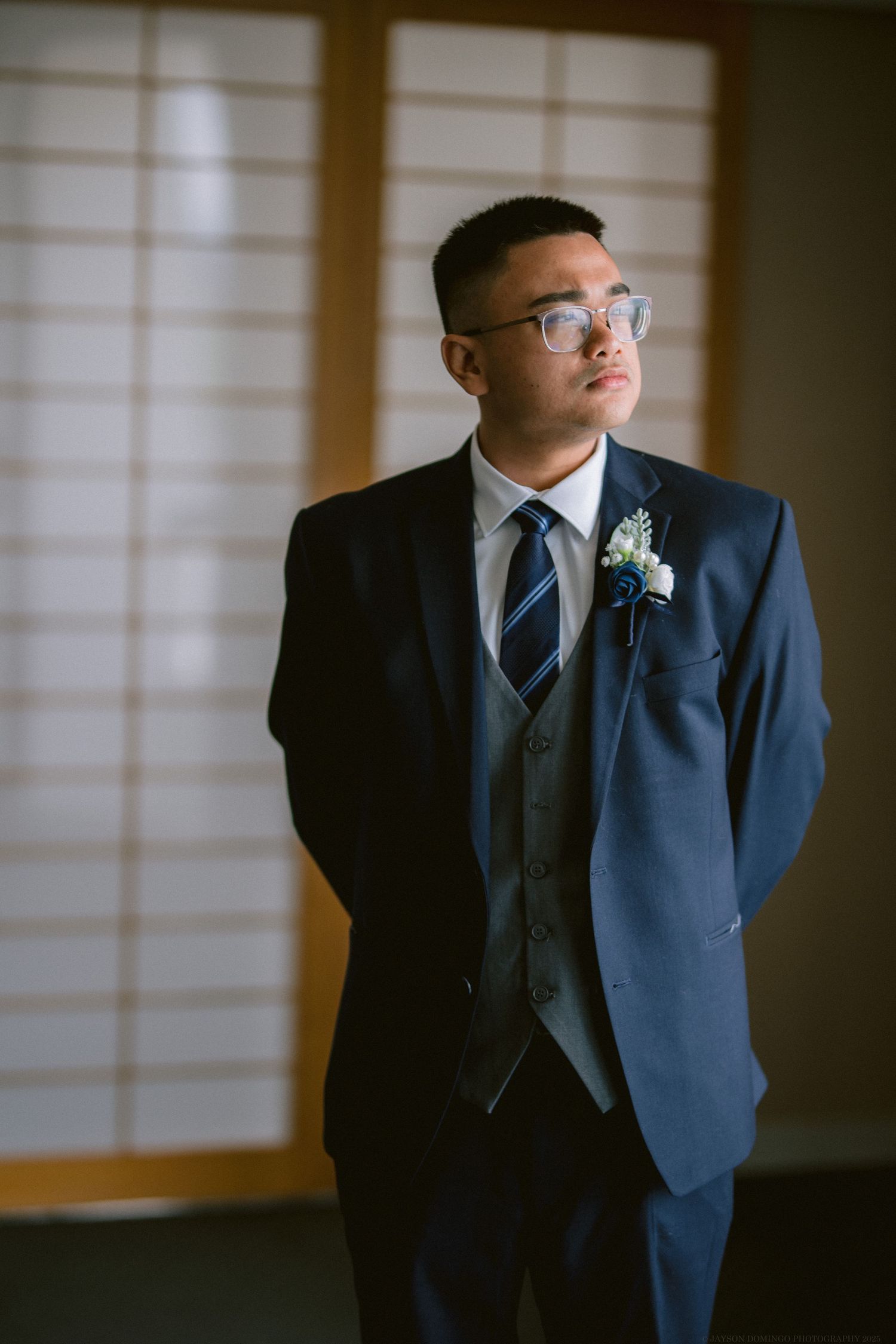 A formal groom in a navy blue three-piece suit and striped tie stands in front of traditional Japanese shoji screens.