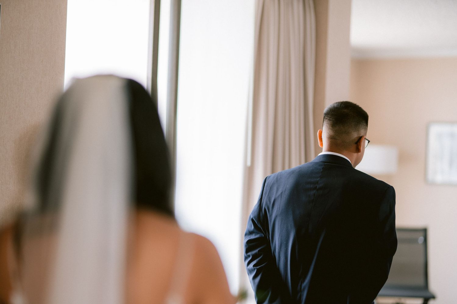 Back view of a groom in a navy suit standing by a window on his wedding day.