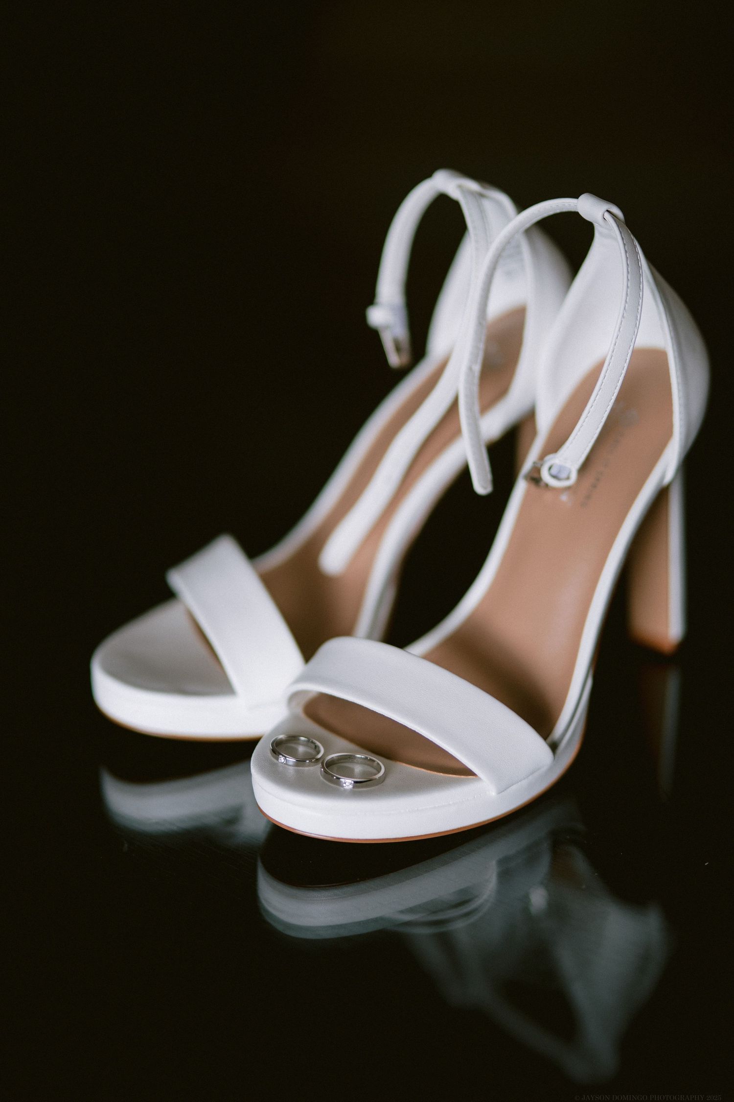 Elegant white bridal heels with ankle straps displayed on reflective surface.