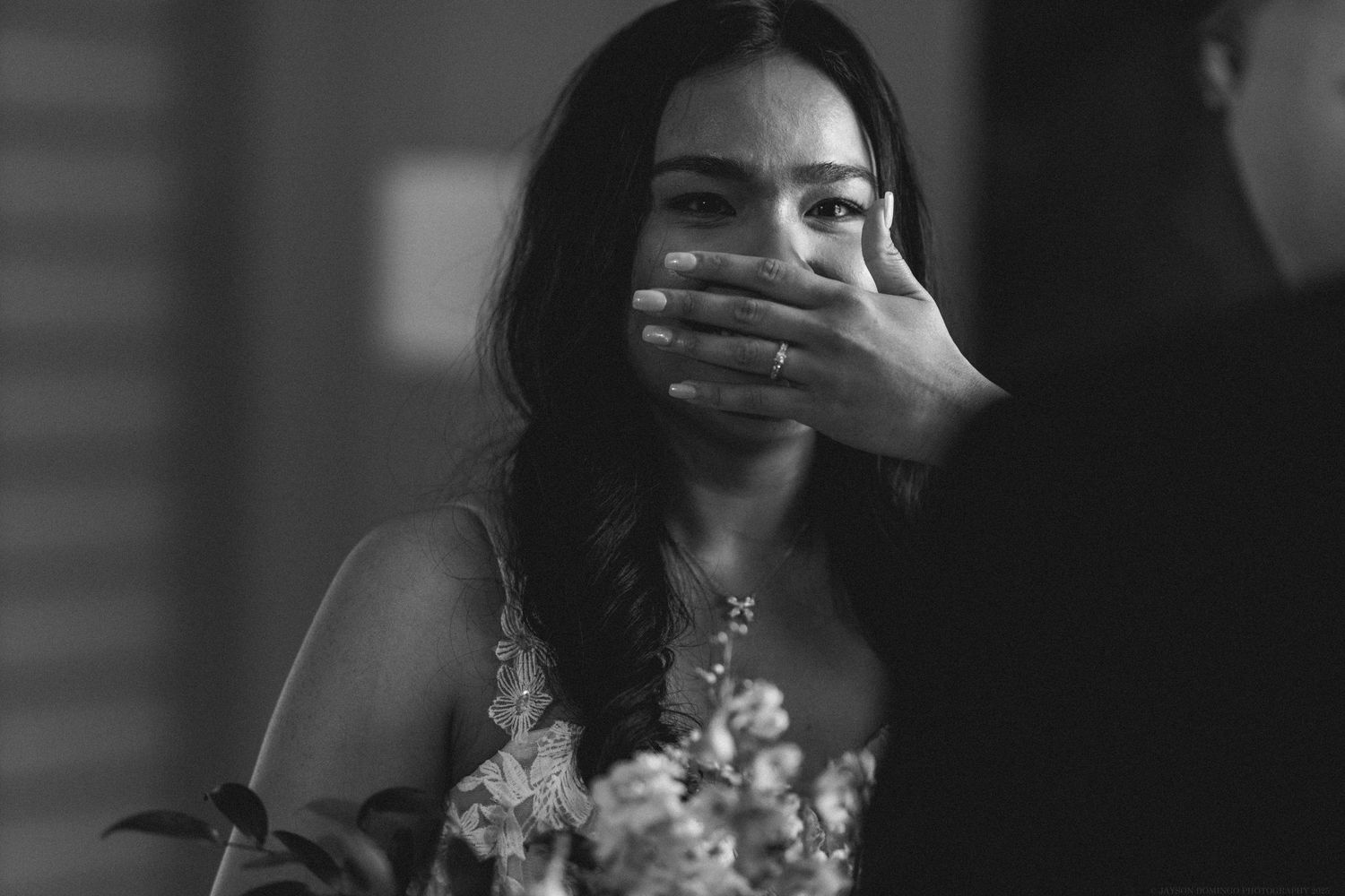 Emotional black and white portrait of a bride in a lace dress covering her mouth.