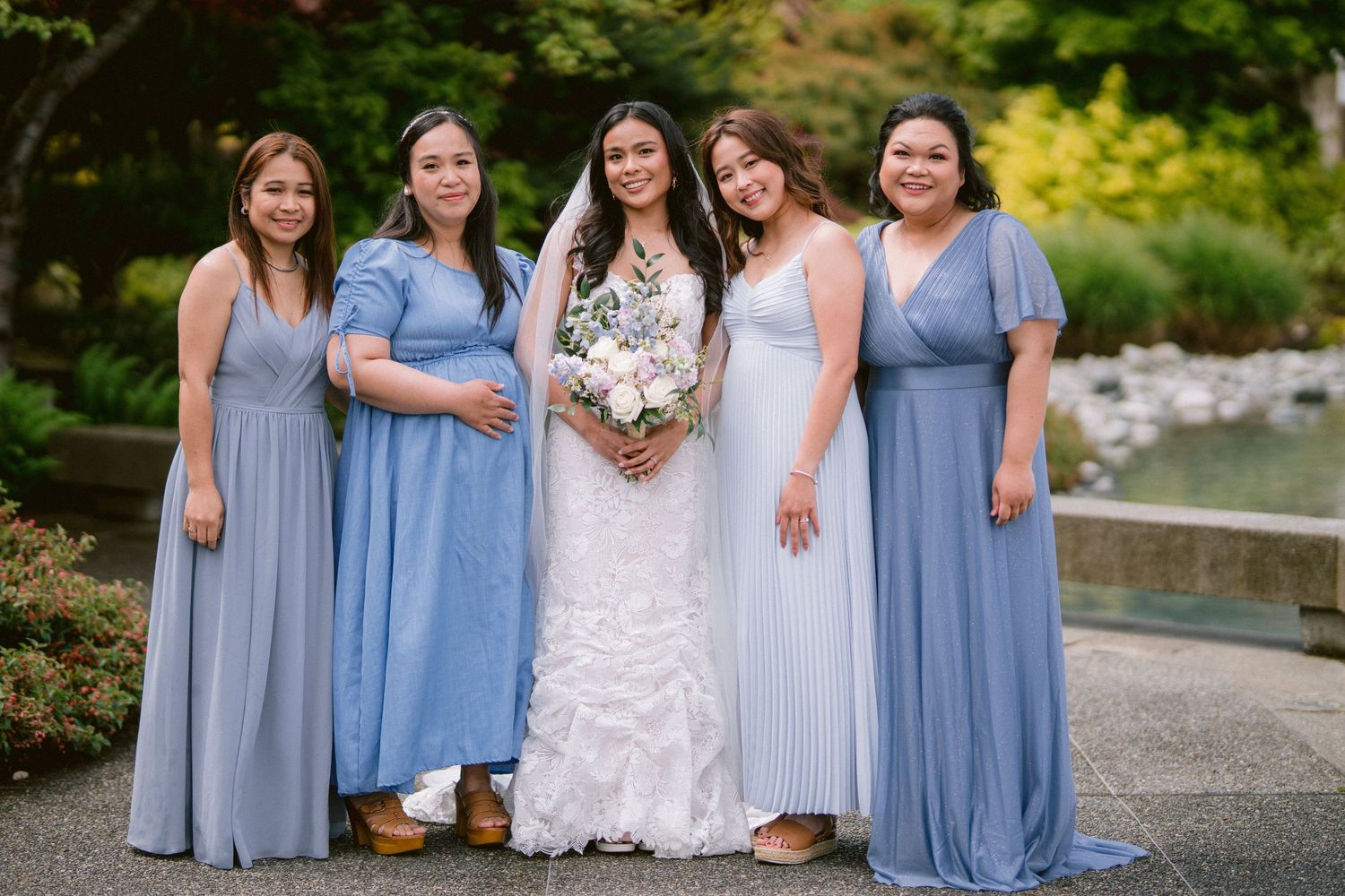 Bridal party in coordinating blue and gray dresses poses outdoors with bouquets.
