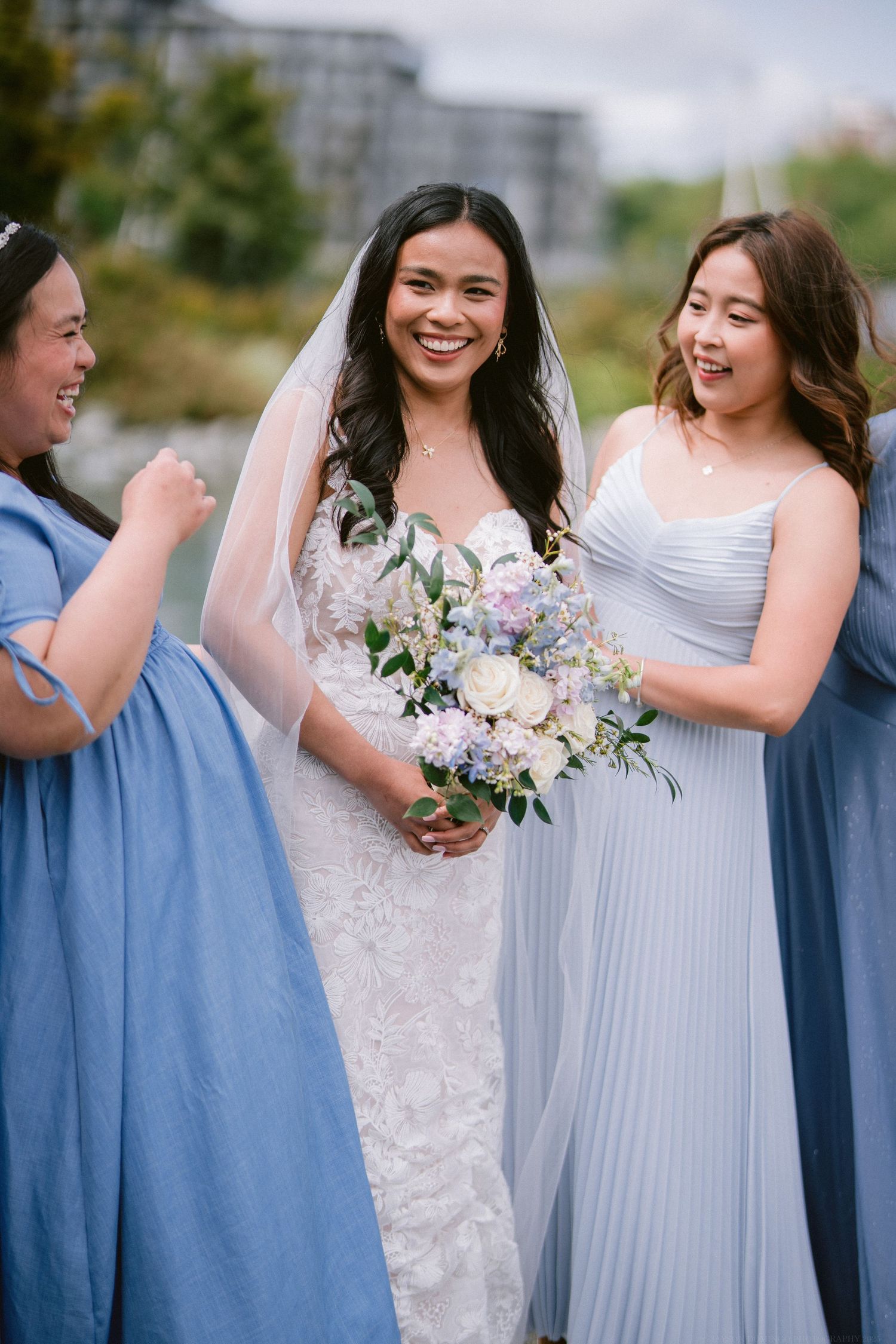 Bridesmaids in flowing blue dresses surround a radiant bride holding a pastel bouquet on her wedding day.