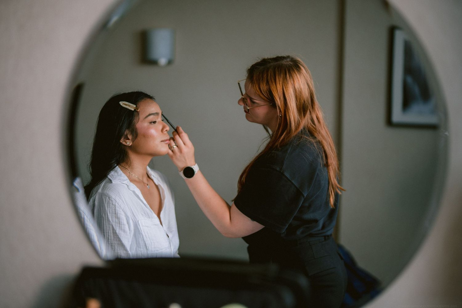 A makeup artist applying cosmetics during bridal preparation in a circular mirror.