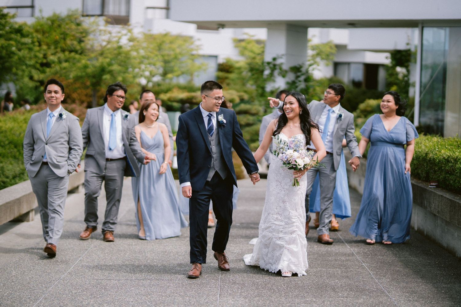 Wedding party in coordinated blue and grey attire walks outdoors along modern building path.