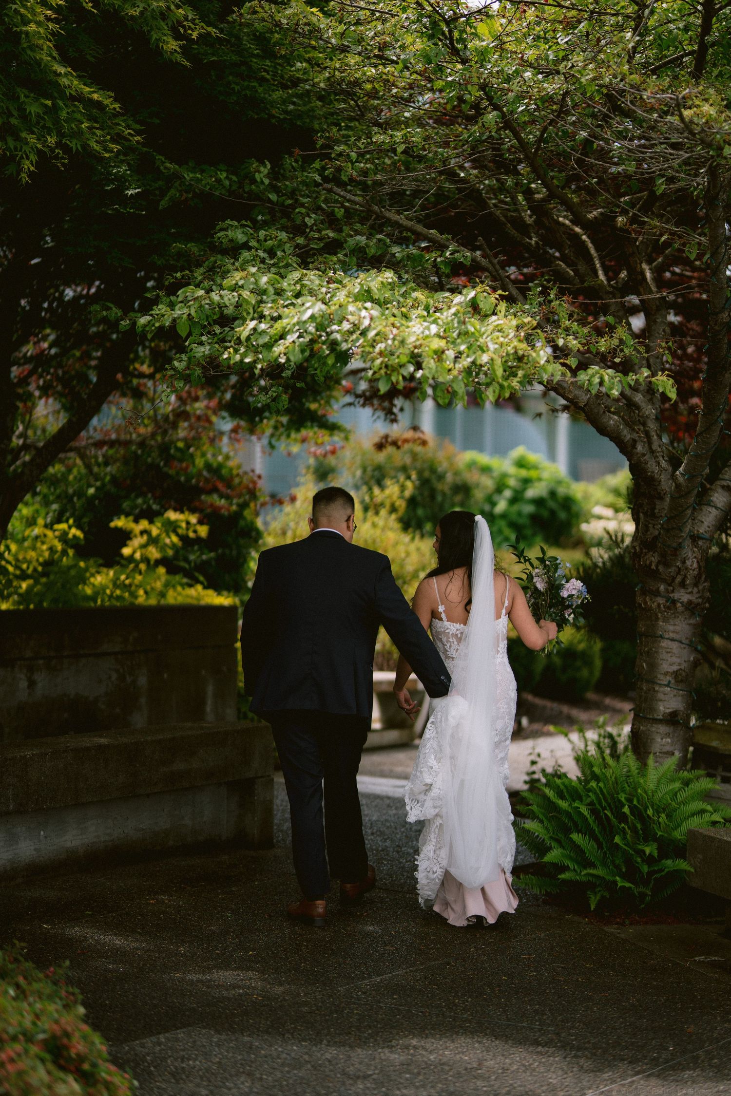 Newlyweds walk down a garden path under lush green trees after their wedding ceremony.
