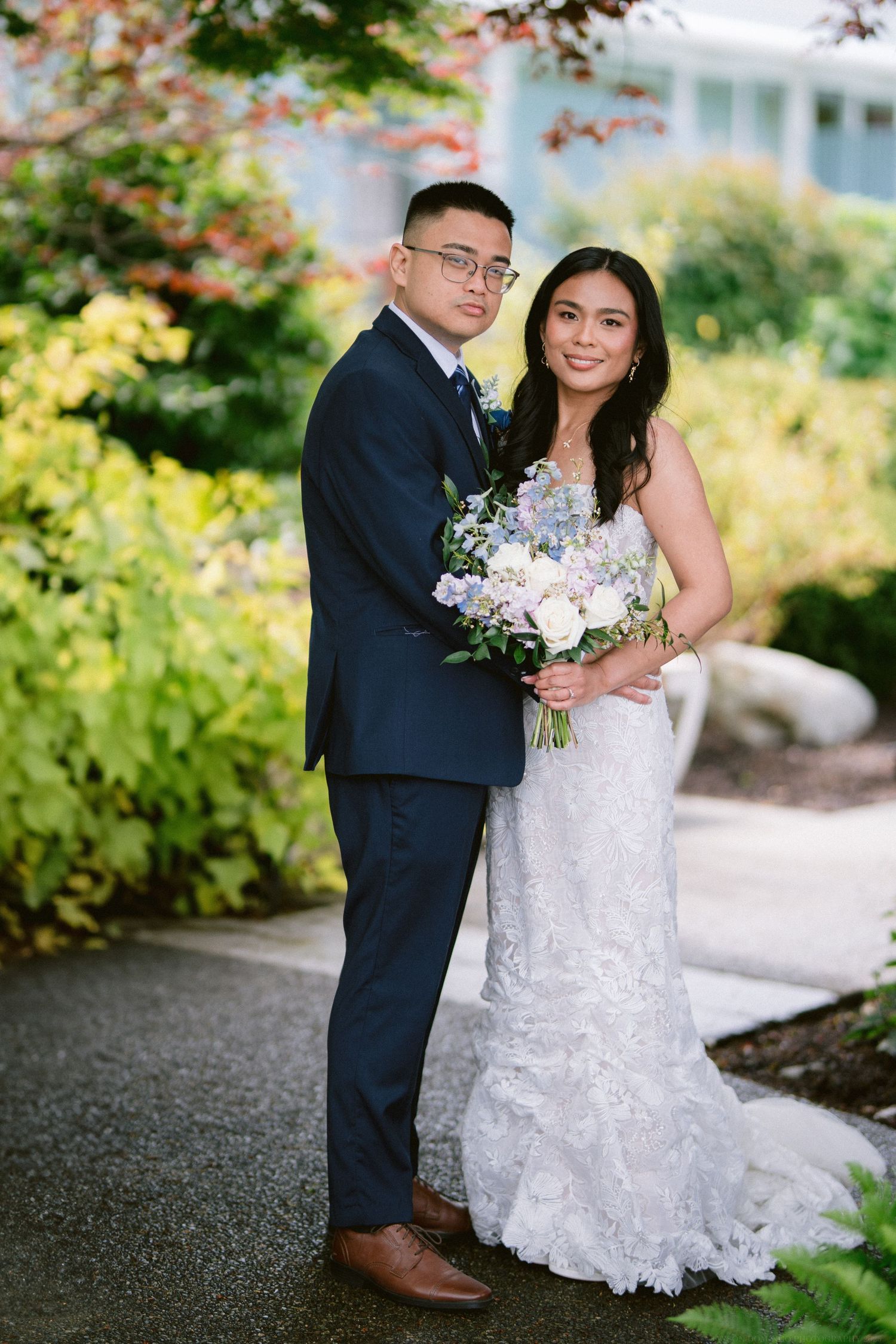 A couple poses in a garden setting on their wedding day, with the bride holding a purple and white bouquet.