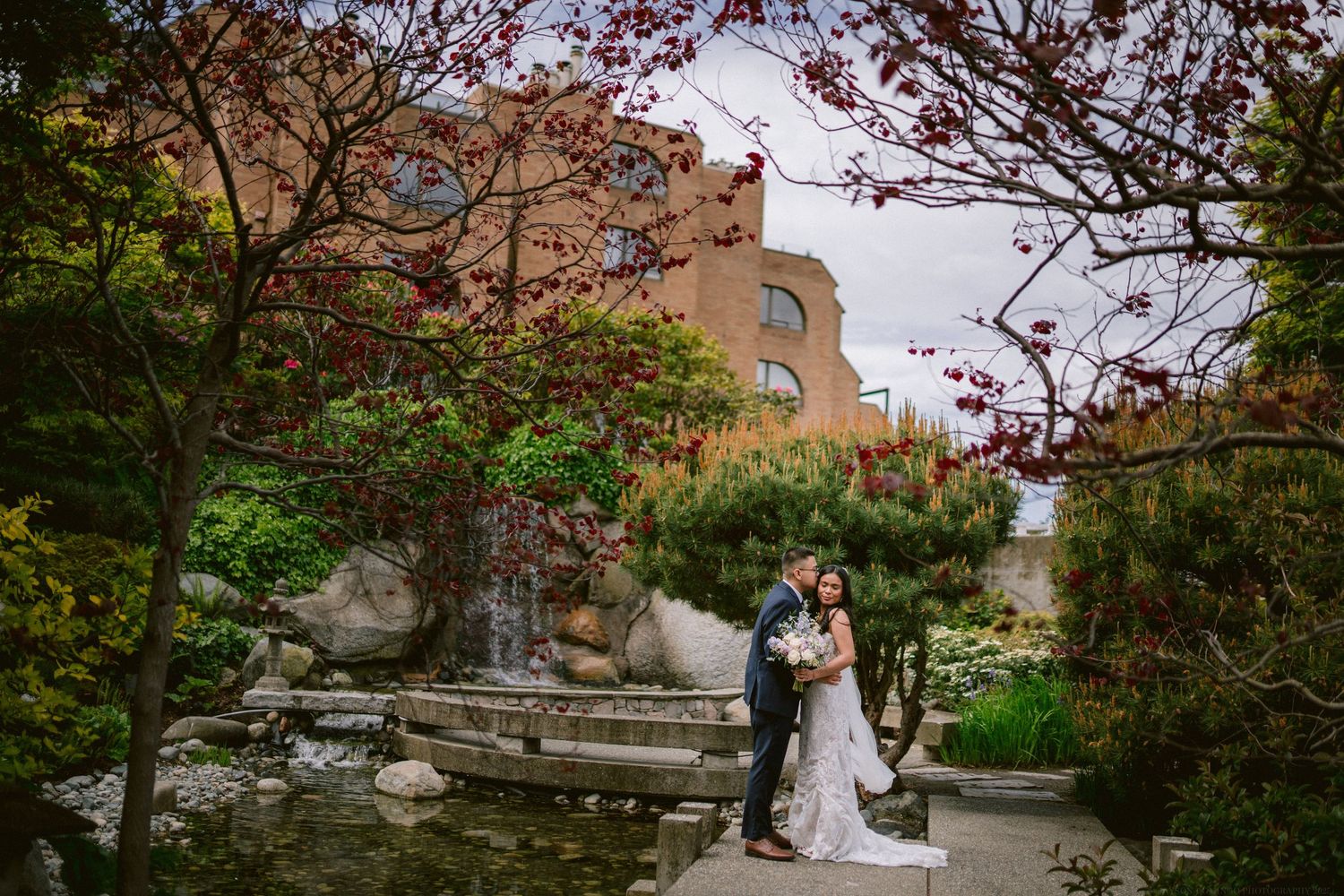 Newlyweds pose for portraits in a garden with brick buildings and cherry blossoms.
