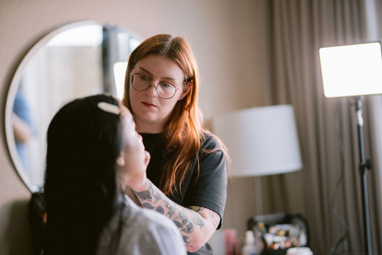 A stylist works on hair and makeup preparations in a hotel room with a round mirror visible.