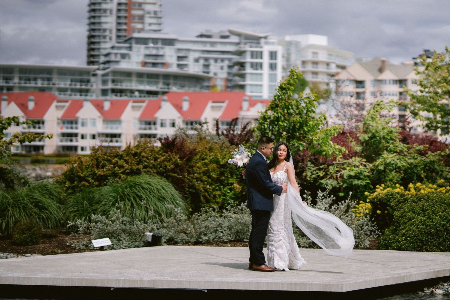 A newlywed couple poses on a deck with city buildings in the background.